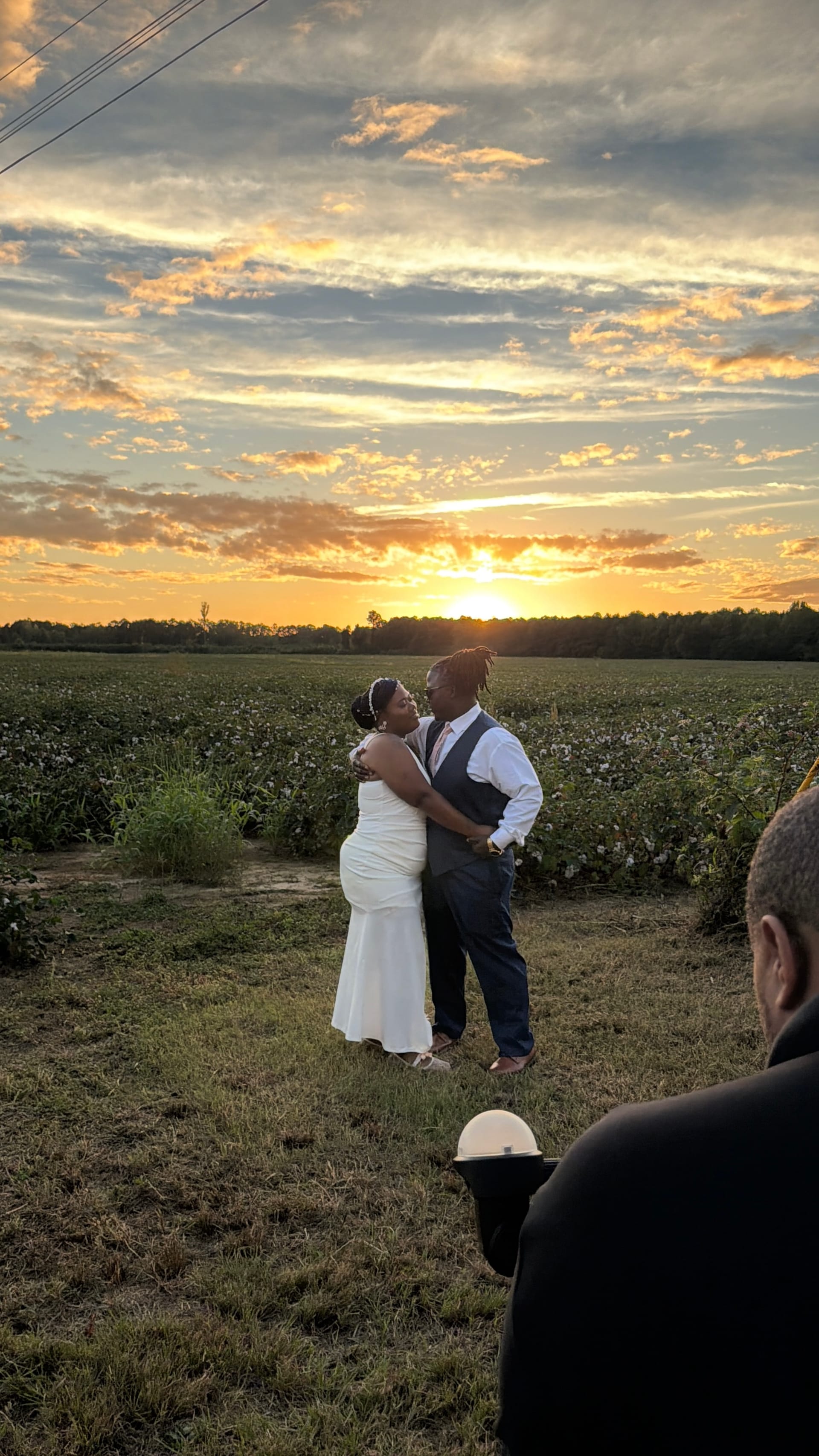 Bride in white gown and groom in grey vest share a romantic embrace in a field at golden hour with a vivid sunset sky glowing behind them at Scarborough House estate.