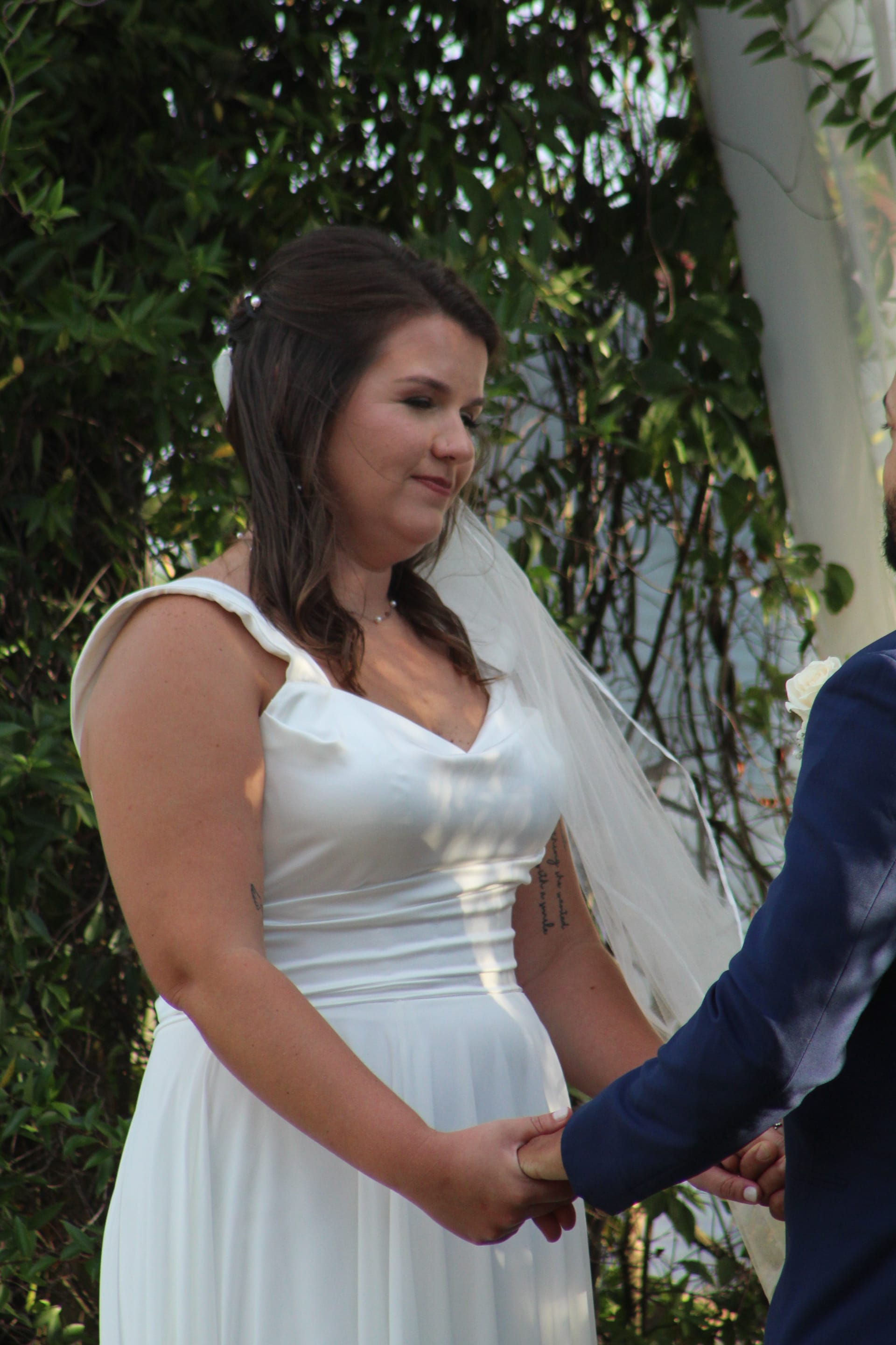 Bride in a white satin gown and veil holds hands with her groom during an outdoor wedding ceremony surrounded by lush green foliage.