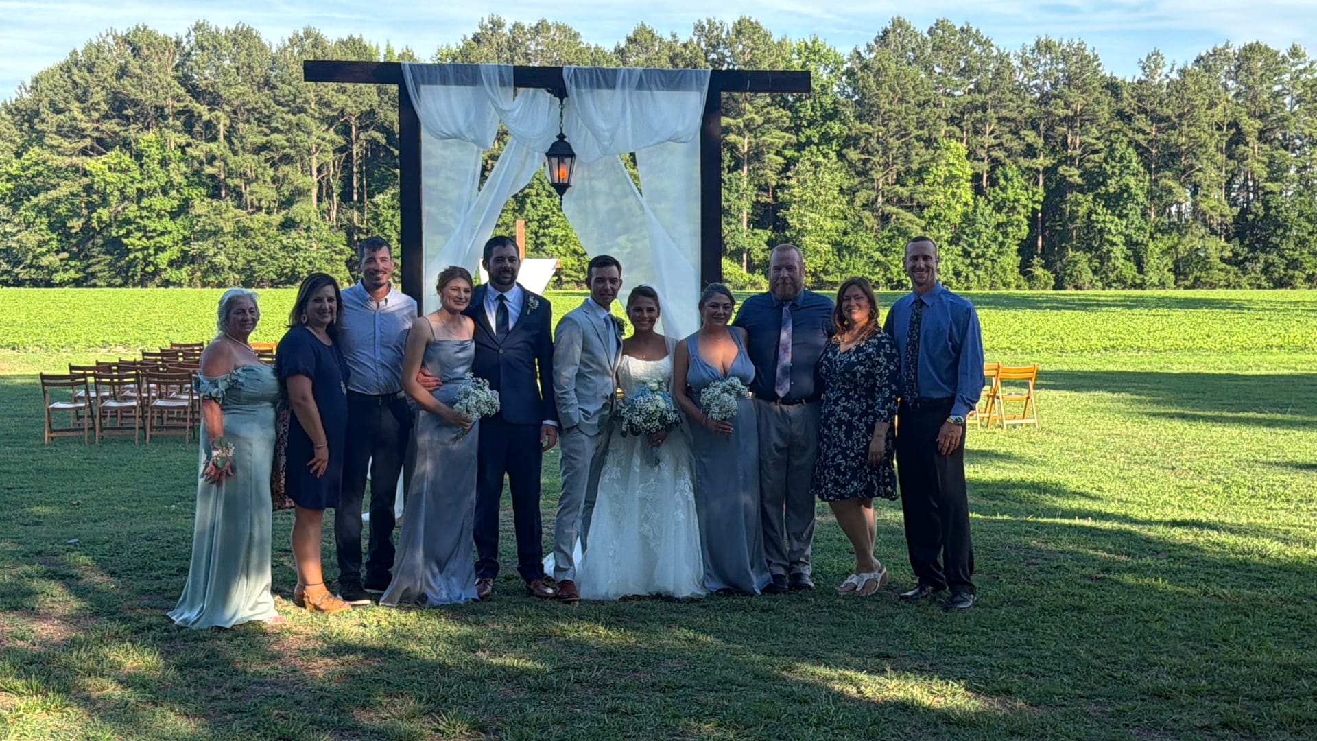Bride and groom stand with their wedding party and family in front of a draped wooden ceremony arch on the sun-lit ceremony lawn at Scarborough House, with lush green farmland fields and pine trees in the background.