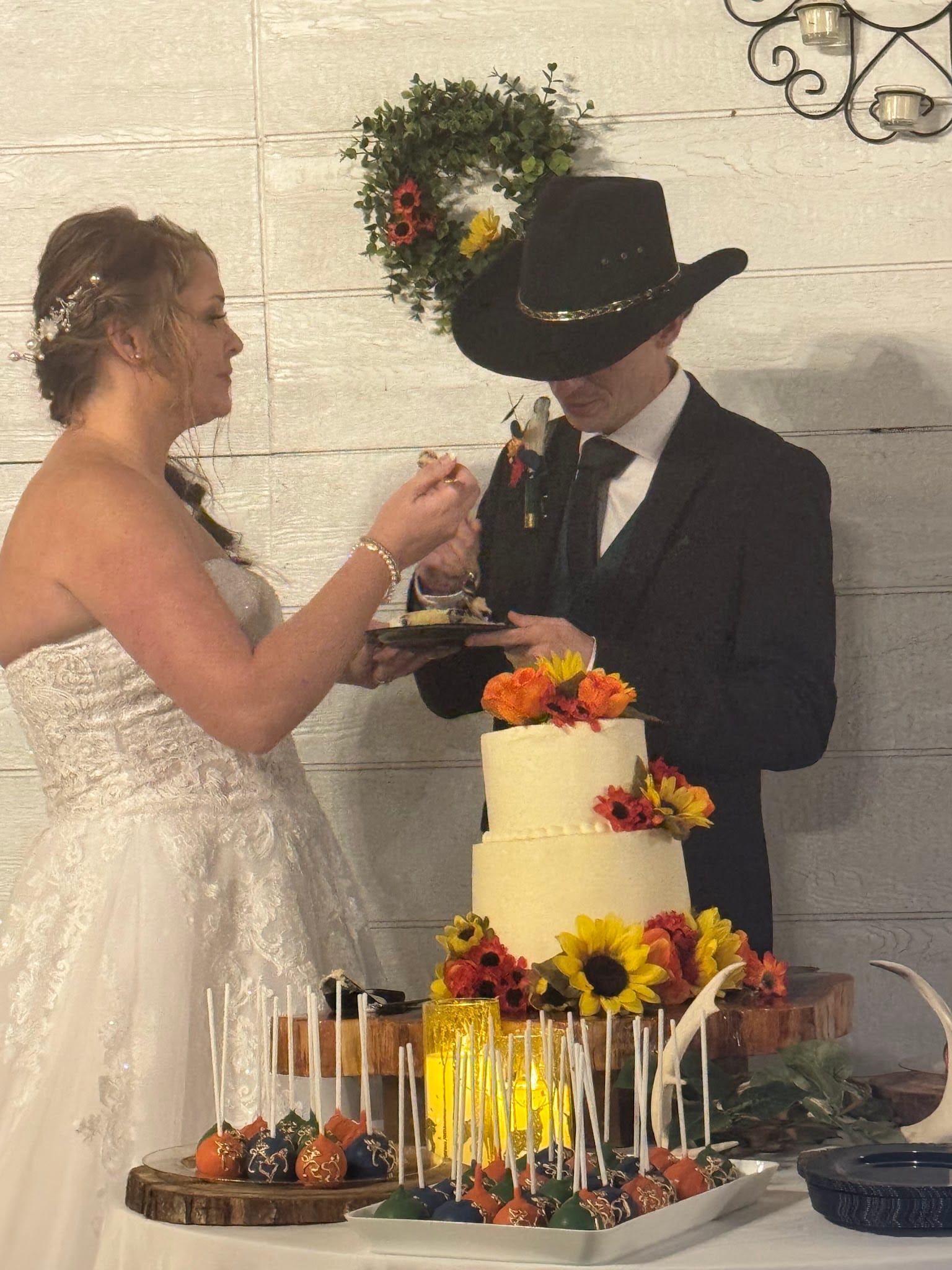 Bride in lace gown feeds cake to groom in black cowboy hat beside a three-tier white wedding cake adorned with sunflowers and autumn blooms at a rustic estate reception.