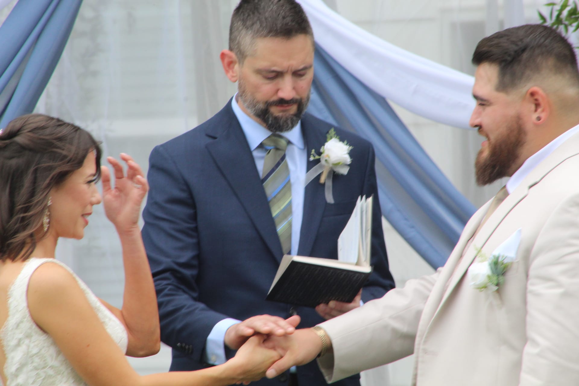 Bride and groom hold hands during their wedding vow exchange as a bearded officiant in a navy suit reads from a black book beneath soft blue and white draped fabric.