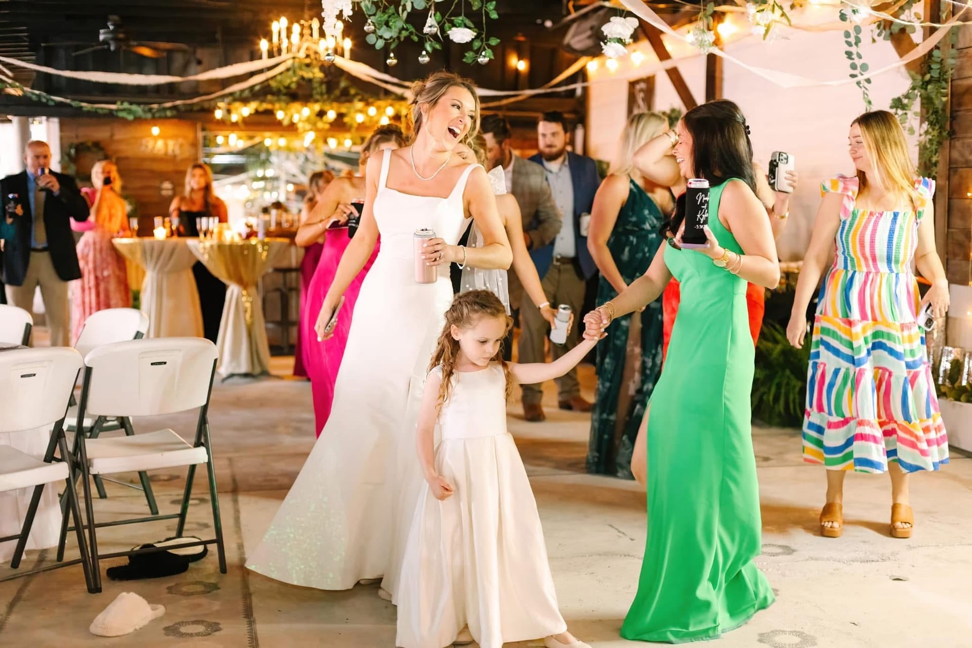 Bride in a white gown laughs while holding hands with a flower girl on the dance floor surrounded by guests at a warmly lit wedding reception.