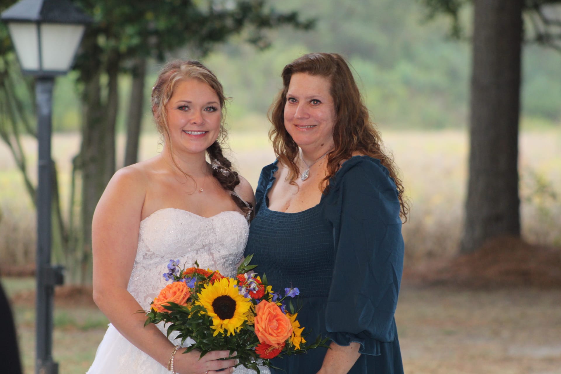 Bride in a strapless white lace gown holding a colorful sunflower and rose bouquet poses smiling alongside a woman in a teal dress beneath the oak trees and open farmland fields at Scarborough House.