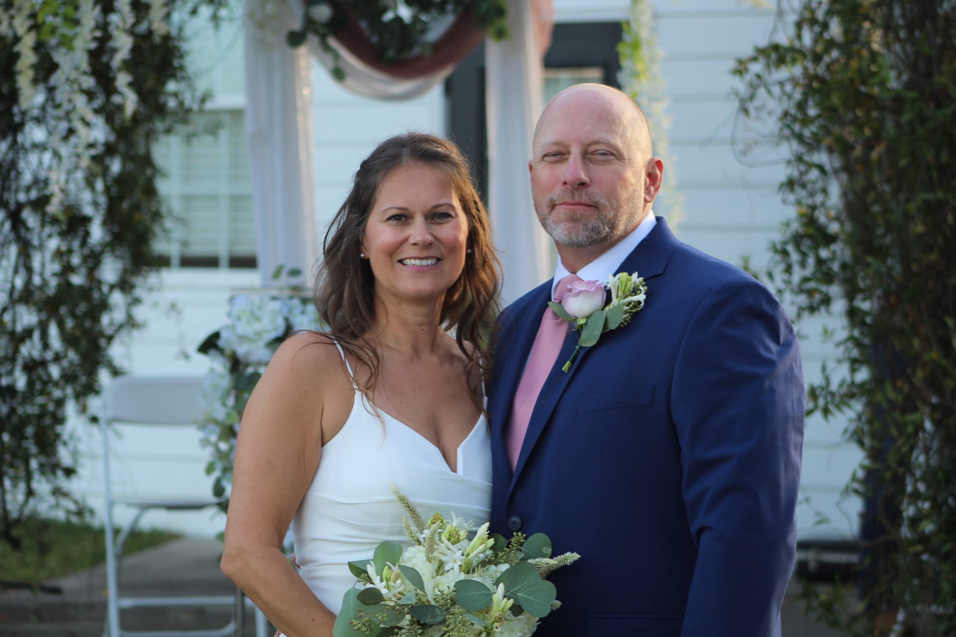 Bride in white gown holding a green and white bouquet stands beside groom in navy suit with pink tie and rose boutonniere in front of the historic Scarborough House manor with lush greenery.