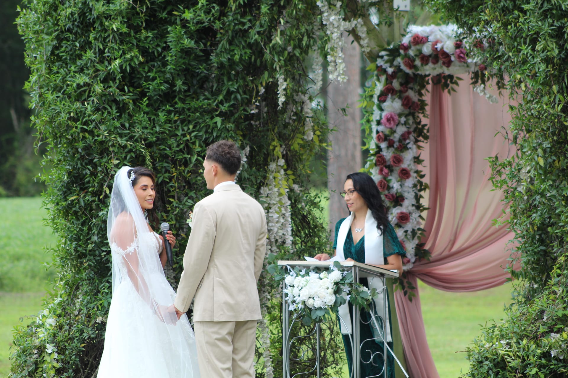 Bride recites her vows to the groom beneath a lush green archway adorned with pink fabric draping and cascading floral arrangements at Scarborough House outdoor ceremony lawn.