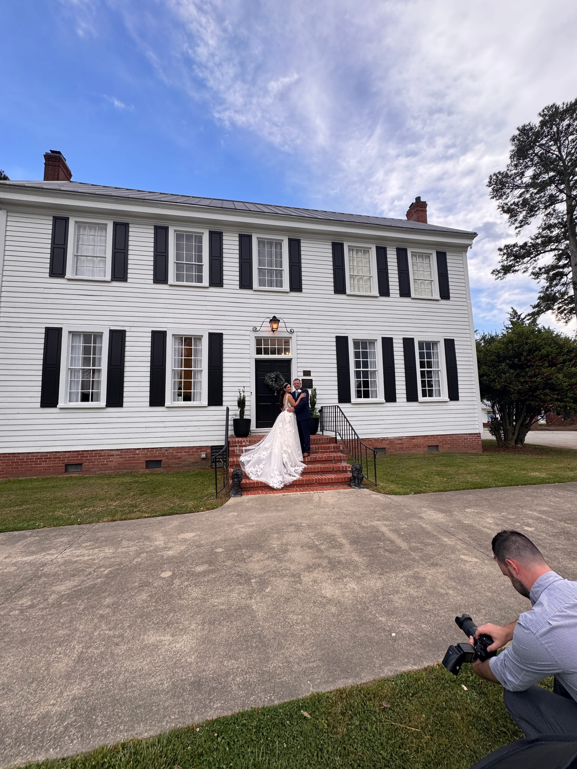 A bride and groom share a romantic embrace on the brick front steps of the historic 1821 Scarborough House manor while a photographer captures the moment from the foreground.