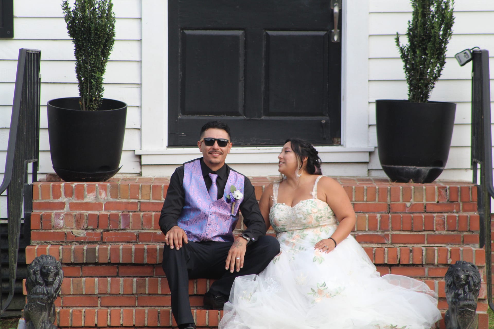 Bride in a floral ball gown and groom in a purple vest and sunglasses sit together on the brick front steps of the historic Scarborough House estate, smiling and sharing a lighthearted moment.