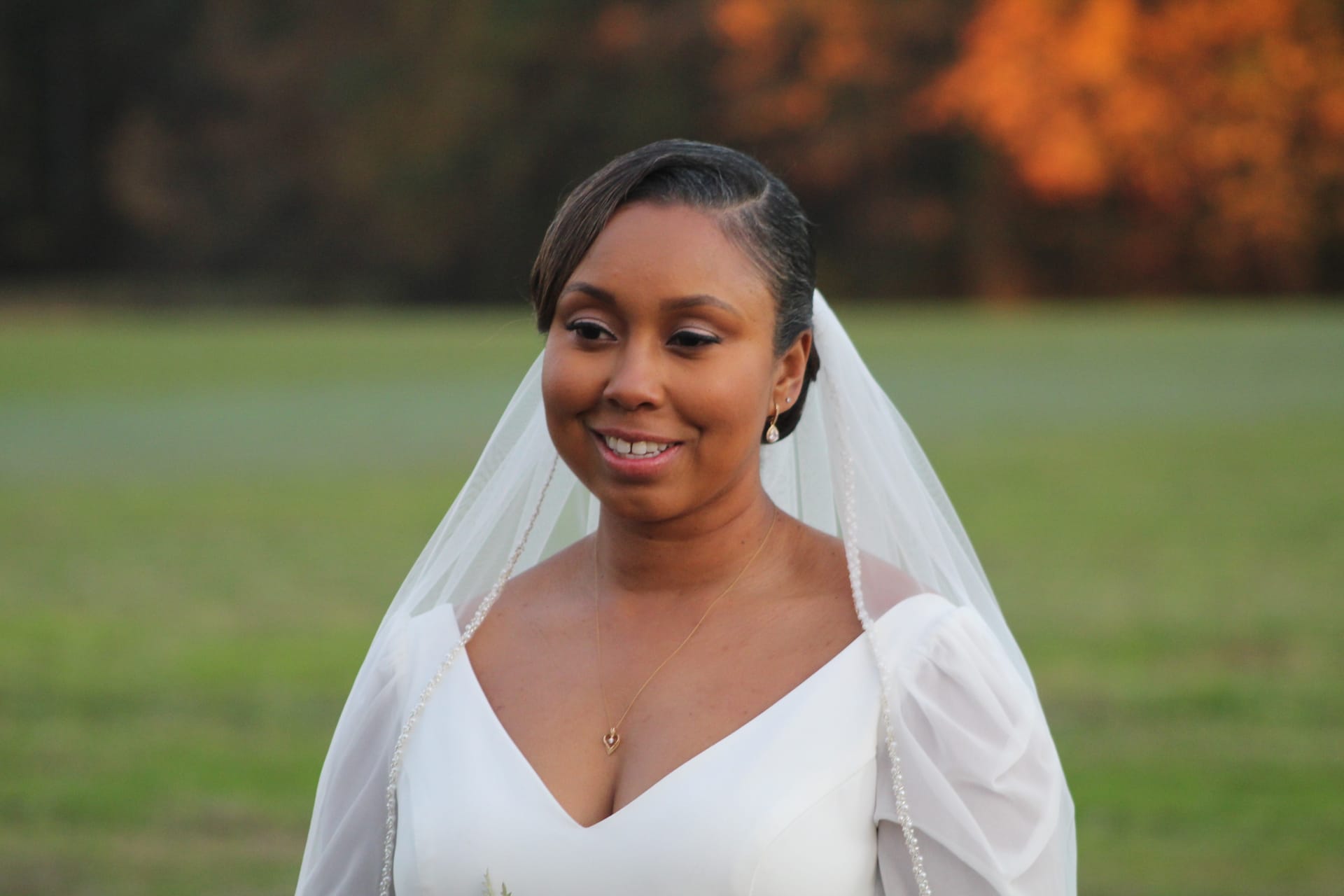 A smiling bride in a white gown and veil stands on the ceremony lawn at Scarborough House with warm golden-hour light and autumn foliage in the background.