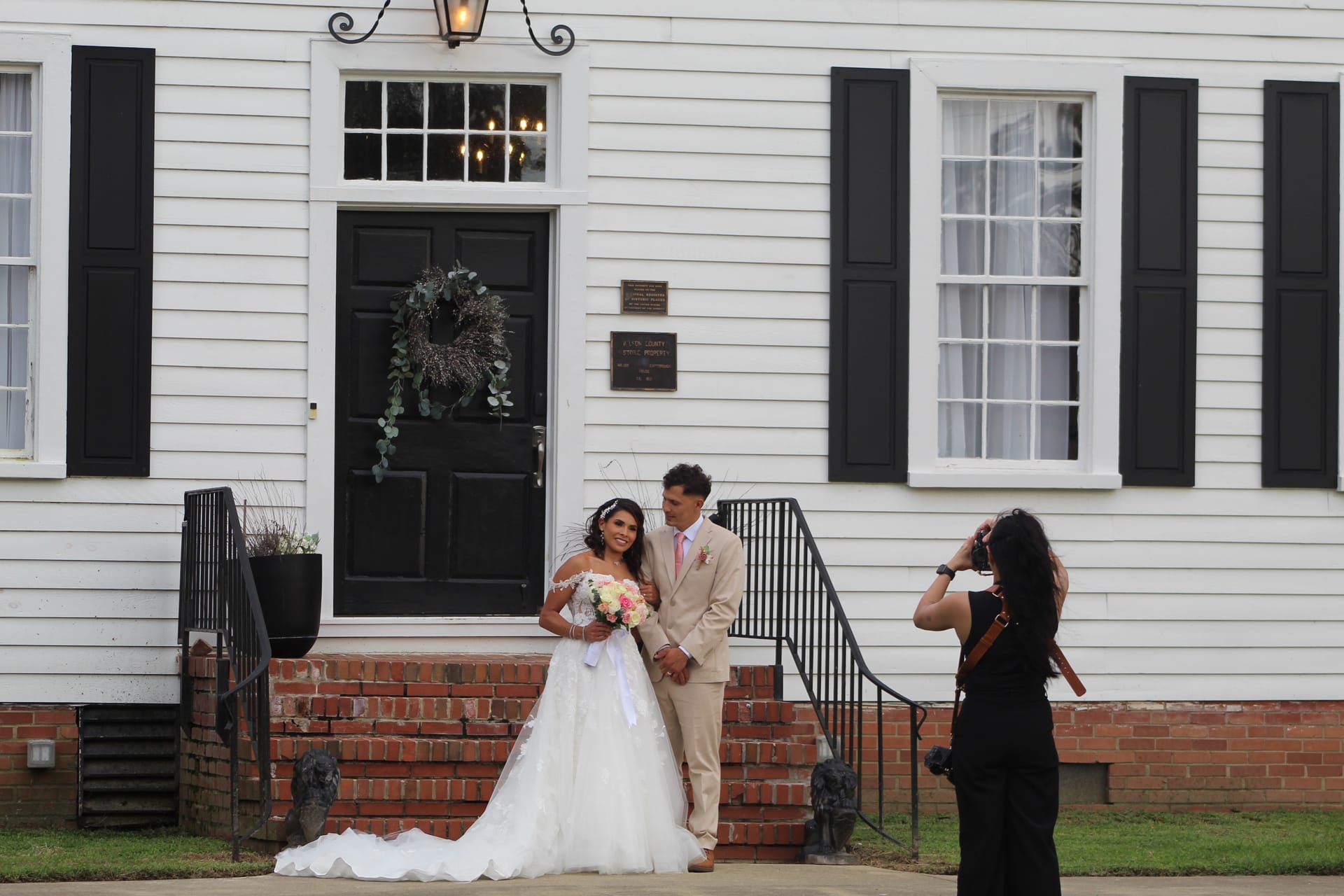 Bride in a white ballgown holding a pink bouquet and groom in a tan suit pose together on the brick front steps of the 1821 Scarborough House manor while a photographer captures their portrait.