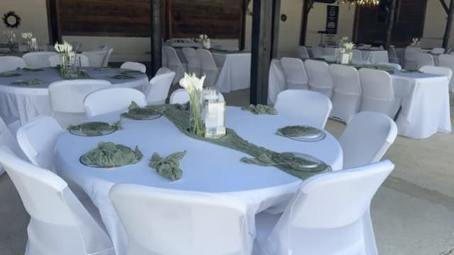 Elegantly set round reception tables with white linens, sage green napkins, silver charger plates, and white calla lily centerpieces inside the rustic grand hall at Scarborough House.