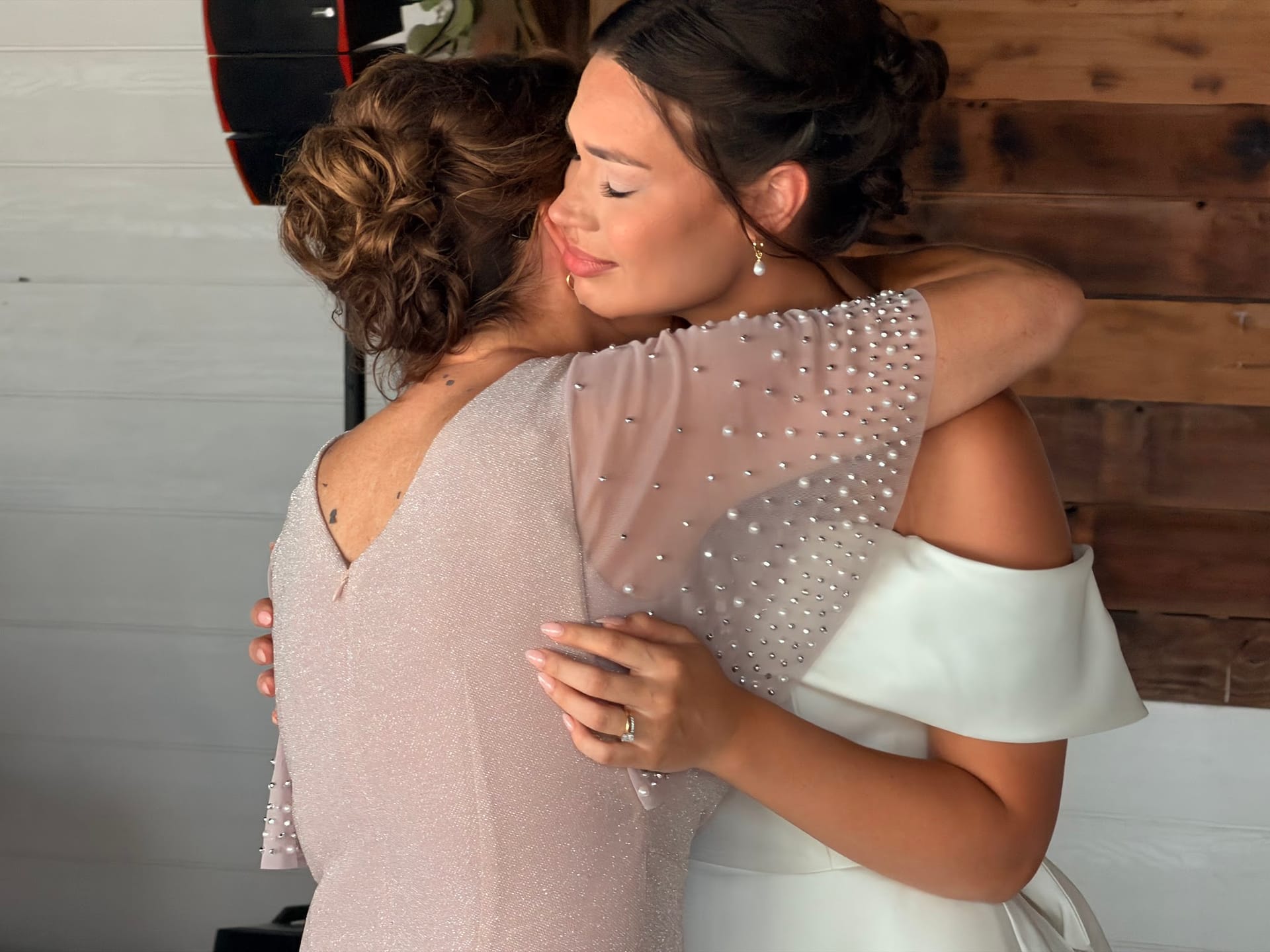 Bride in a white off-shoulder gown shares a heartfelt embrace with a guest in a pearl-embellished blush dress inside a rustic wood-paneled room at Scarborough House.