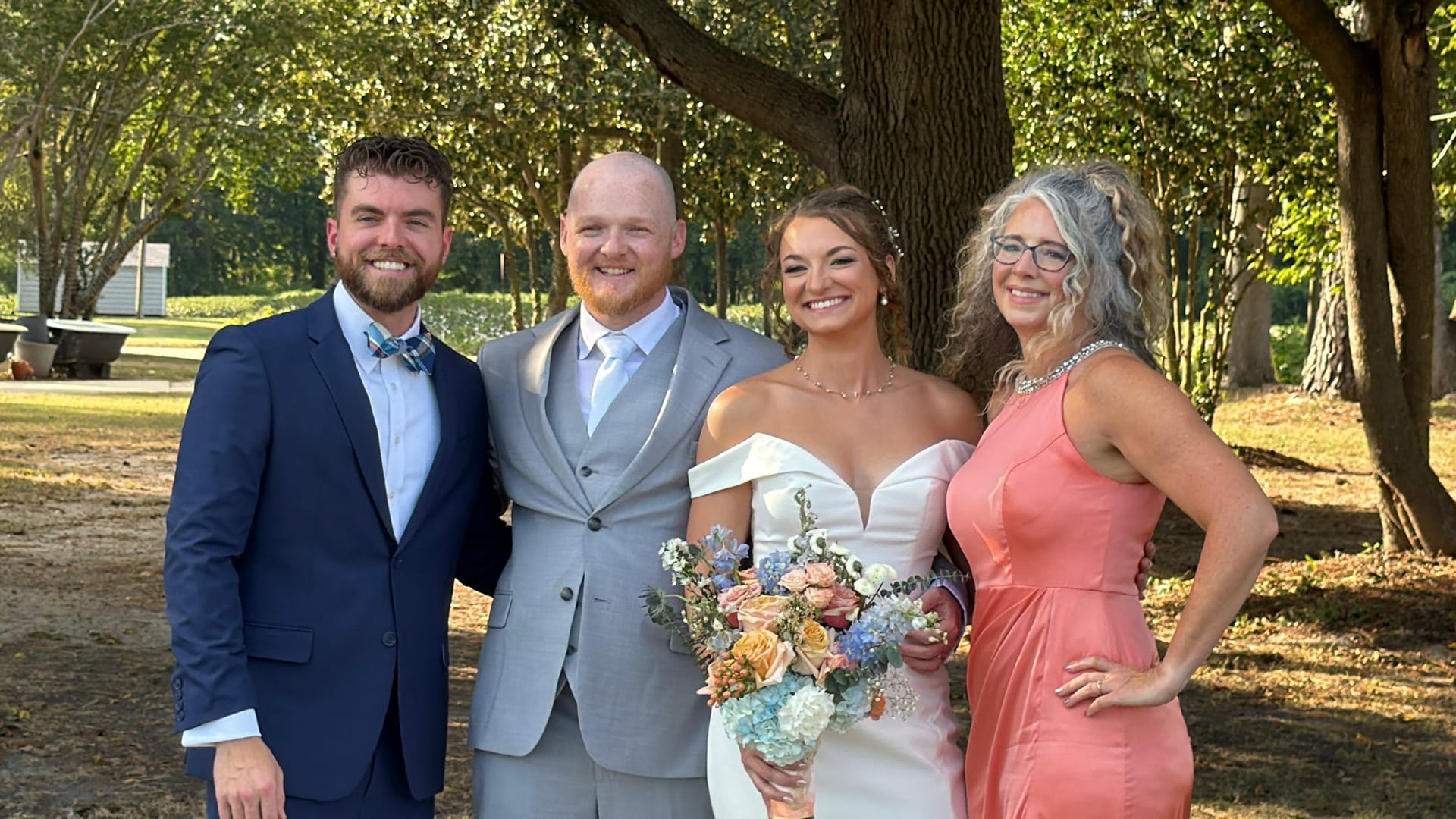 Bride and groom pose with two guests under sunlit oak trees at Scarborough House, the bride holding a pastel floral bouquet of peach roses and blue hydrangeas.