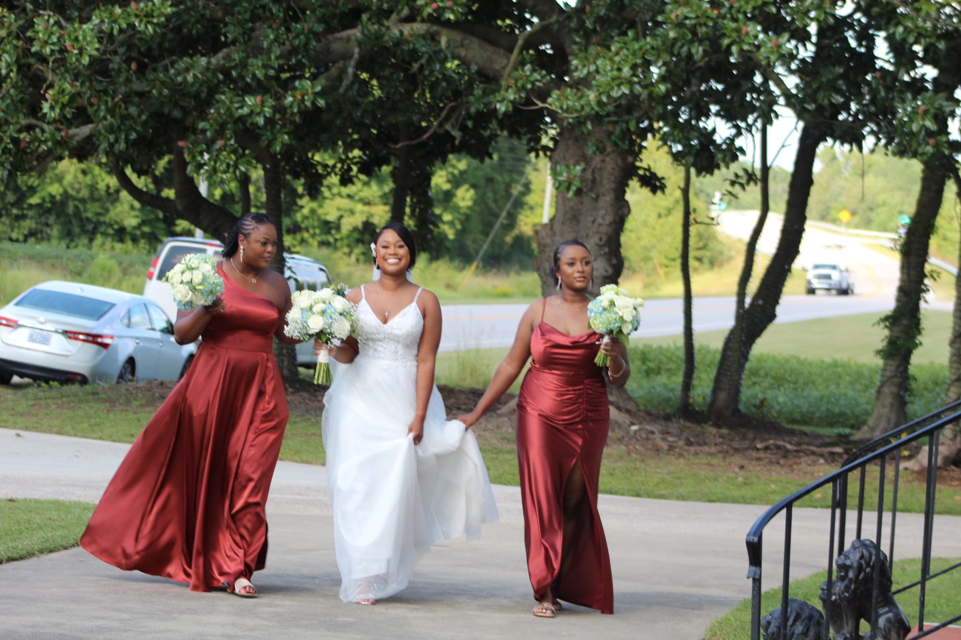 A radiant bride in a white lace gown walks with two bridesmaids in burgundy satin dresses carrying white floral bouquets along the tree-lined driveway of Scarborough House historic estate.