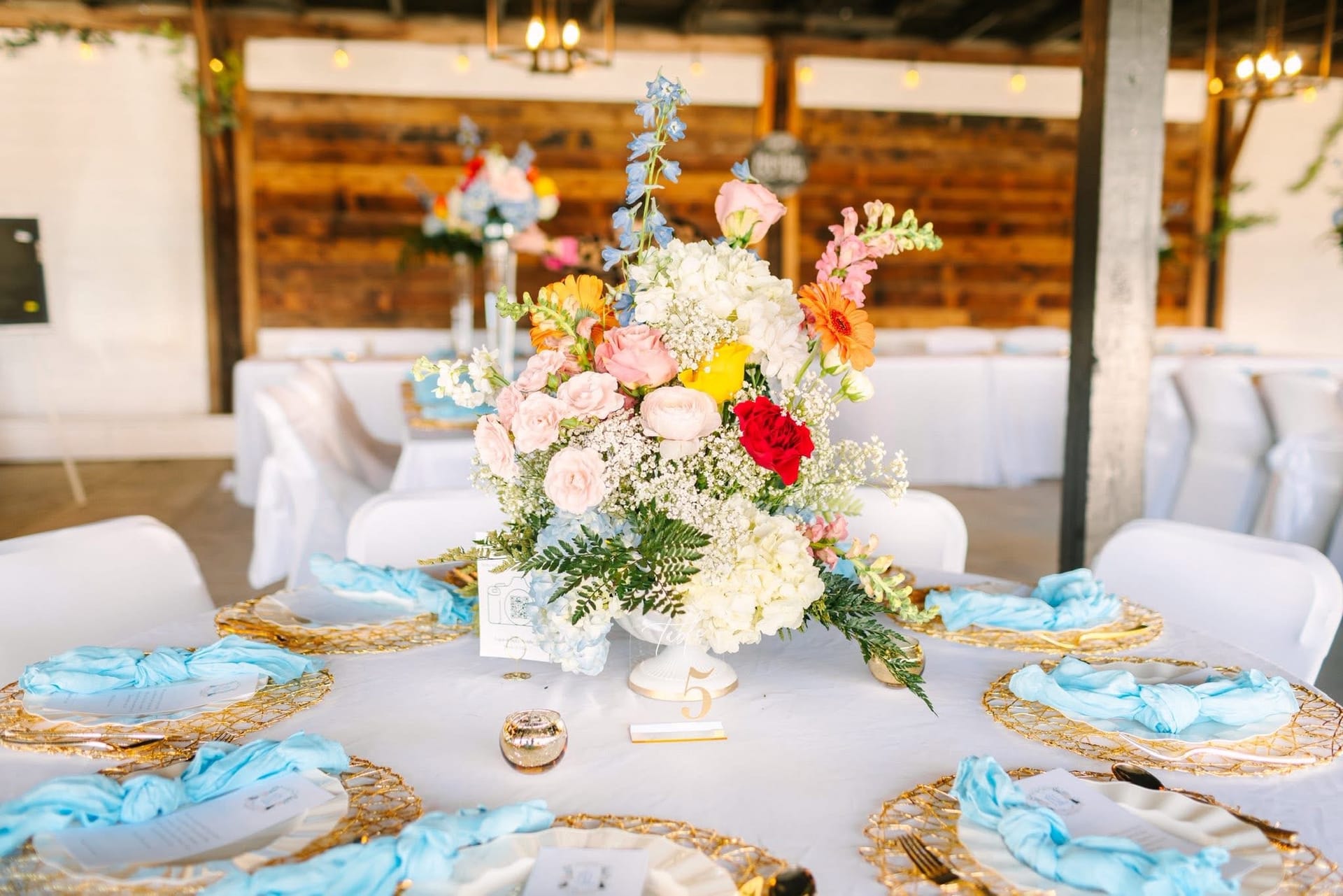 Elegantly styled round reception table at Scarborough House featuring a lush colorful floral centerpiece in a white urn, gold wire charger plates, light blue linen napkins, and a gold table number five.