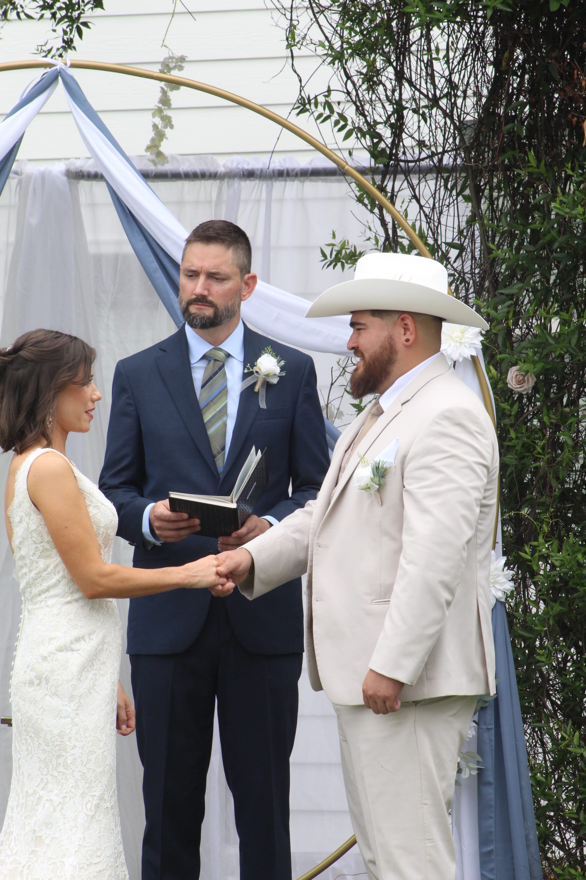 Bride and groom hold hands during their outdoor wedding ceremony beneath a gold circular arch draped with blue and white fabric at Scarborough House estate.