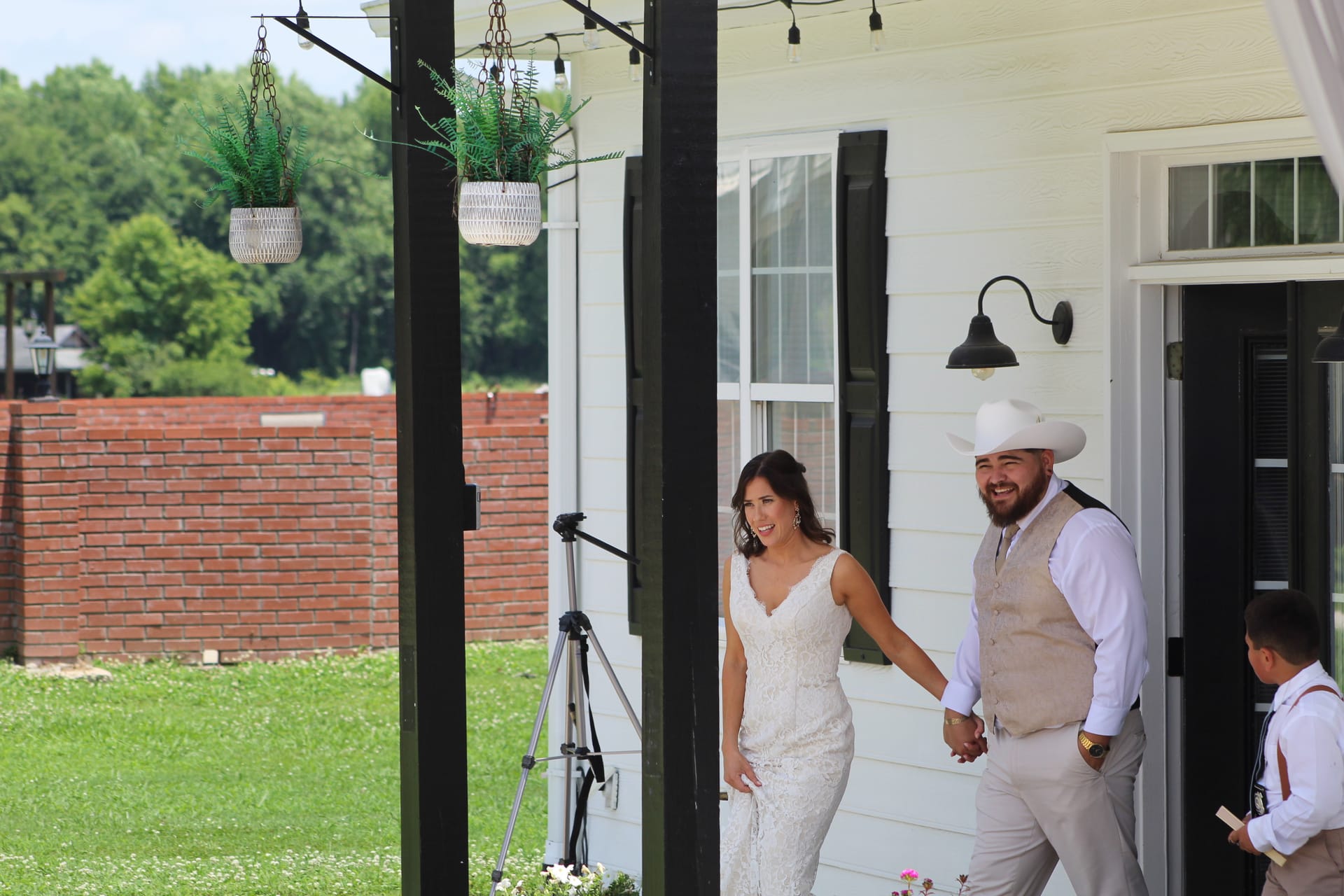 Smiling bride in a lace gown and groom in a tan vest and white cowboy hat walk hand-in-hand out of the Scarborough House porch doorway on their wedding day.