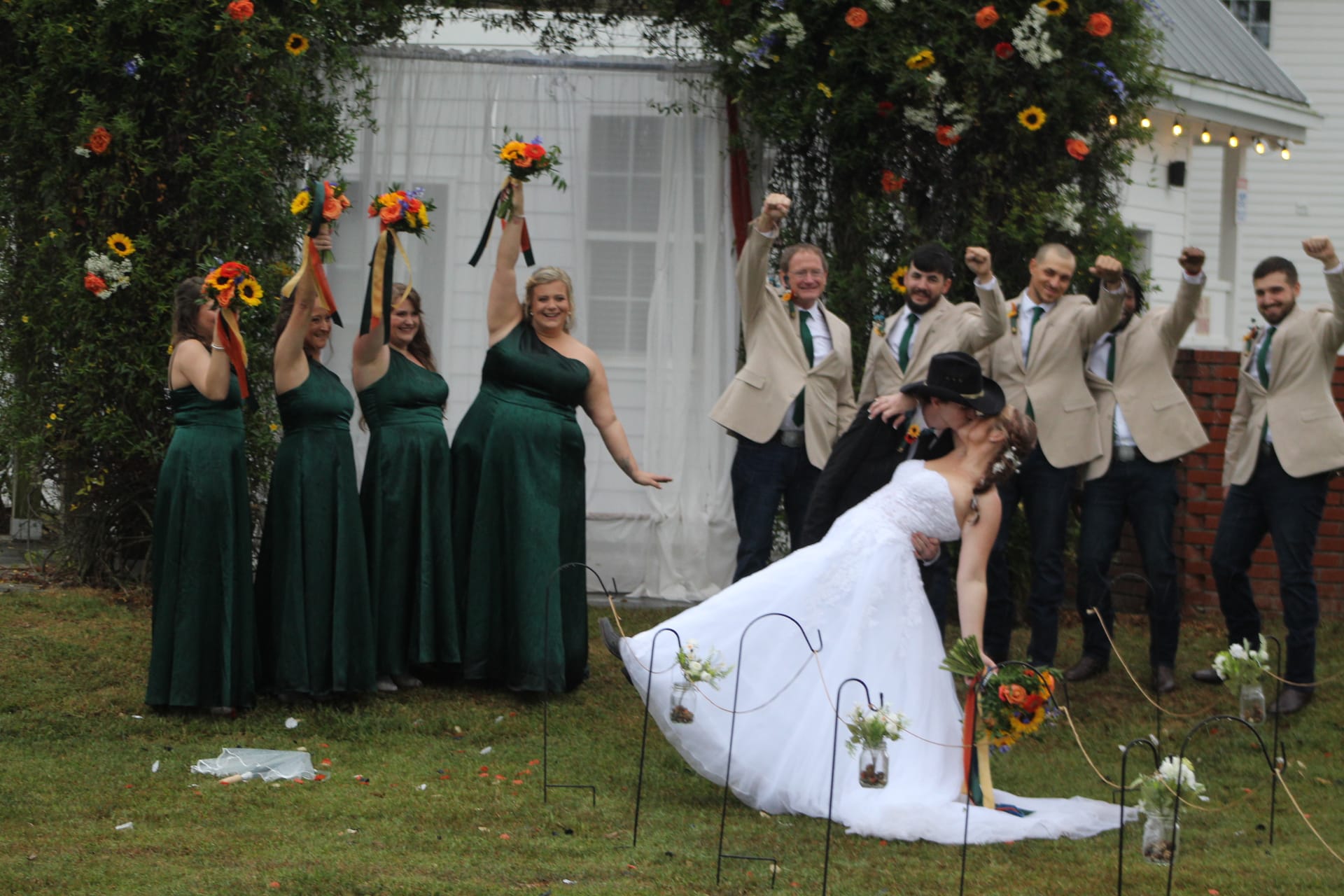 Bride and groom share a dramatic dip kiss on the ceremony lawn as bridesmaids in emerald green gowns raise colorful bouquets and groomsmen in tan blazers cheer beneath a floral arch in front of the historic Scarborough House manor.