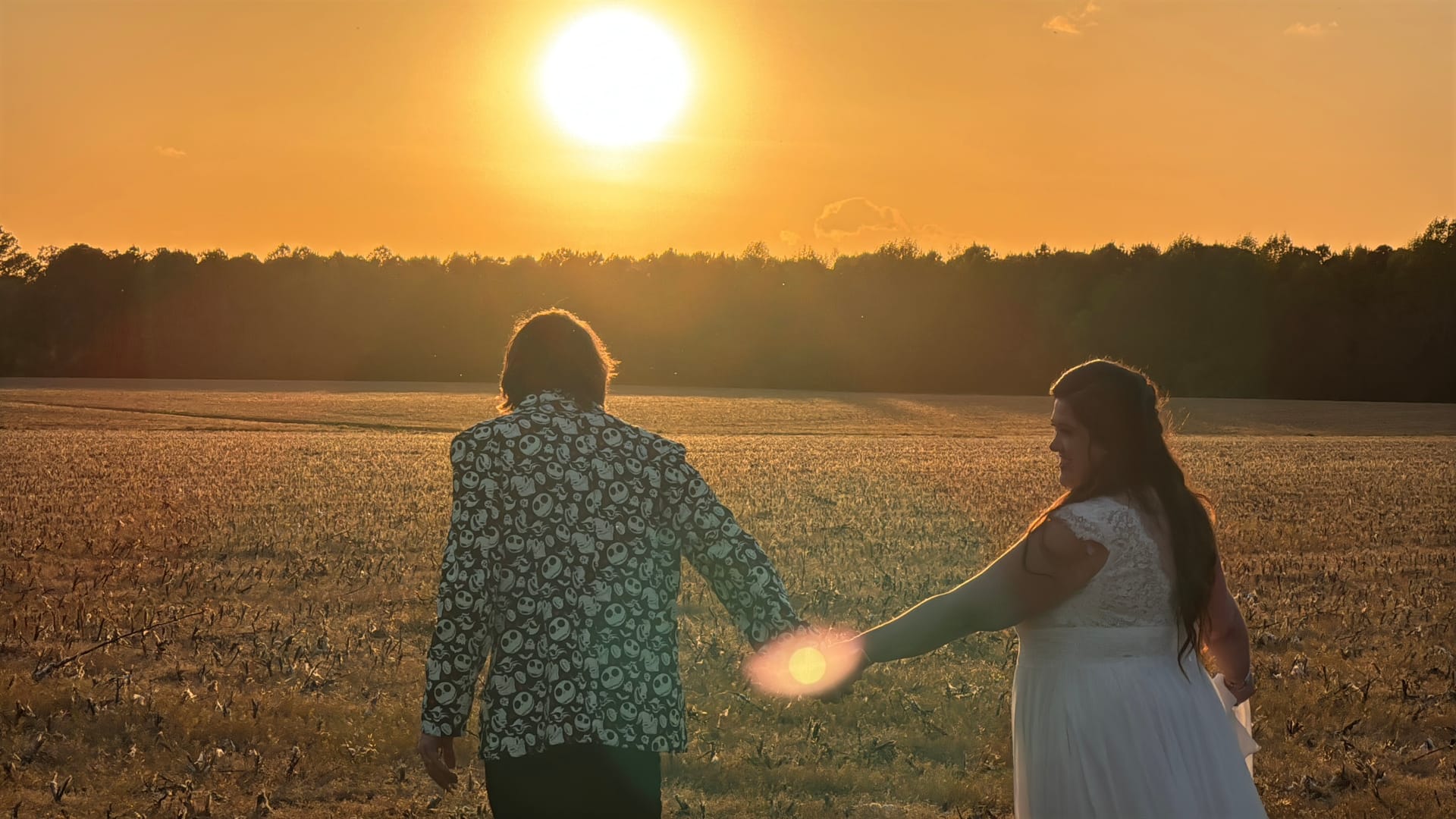Bride in a white lace gown and groom in a patterned jacket walk hand in hand through open farmland fields toward a brilliant golden sunset at Scarborough House estate.