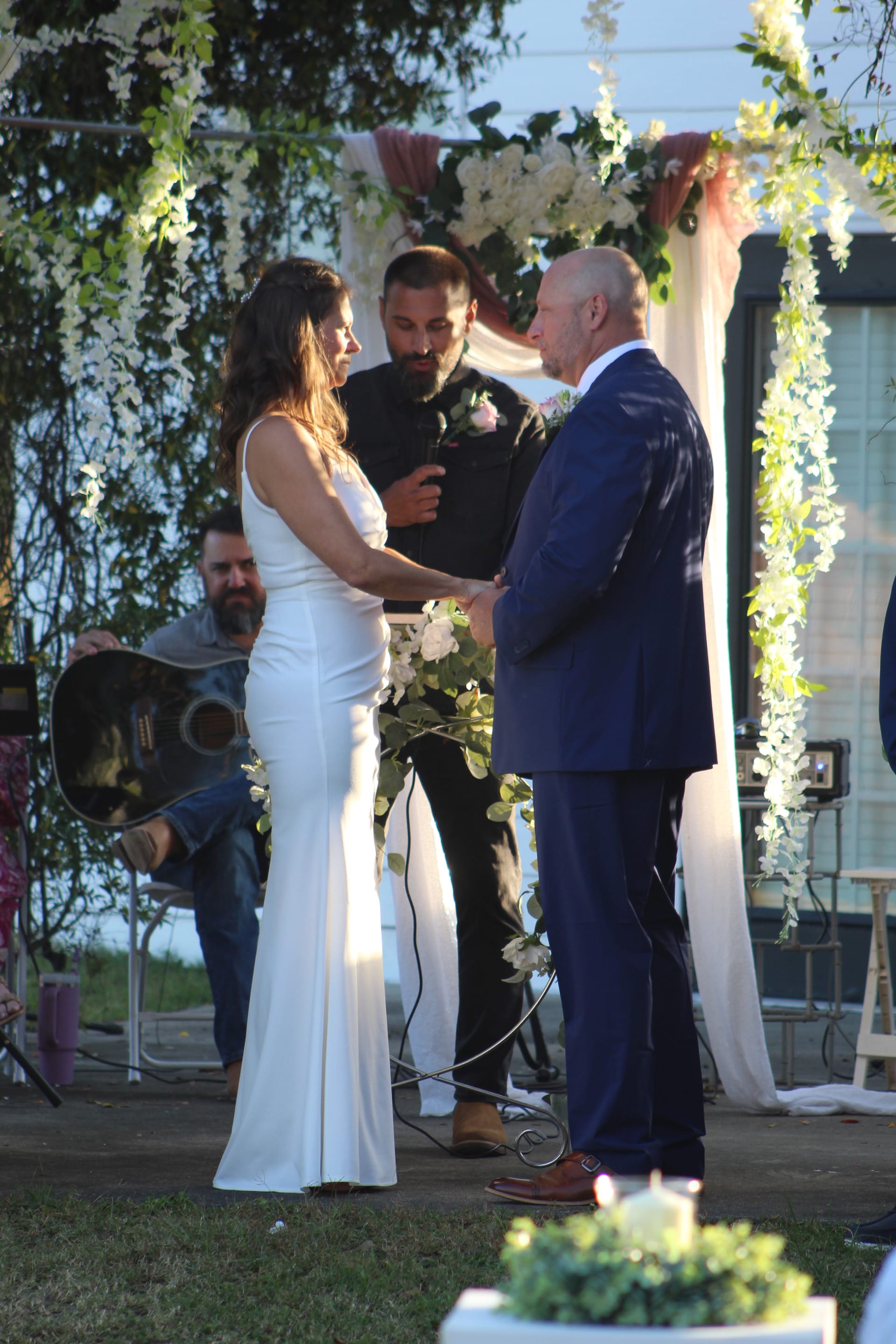 Bride in a white gown and groom in a navy suit exchange vows beneath a lush floral arch decorated with white wisteria and greenery at a historic estate wedding ceremony.