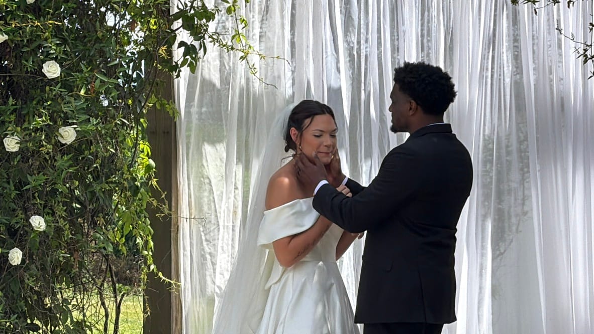 Groom tenderly holds brides face during their outdoor wedding ceremony beneath a white draped backdrop adorned with white roses and lush greenery at Scarborough House.