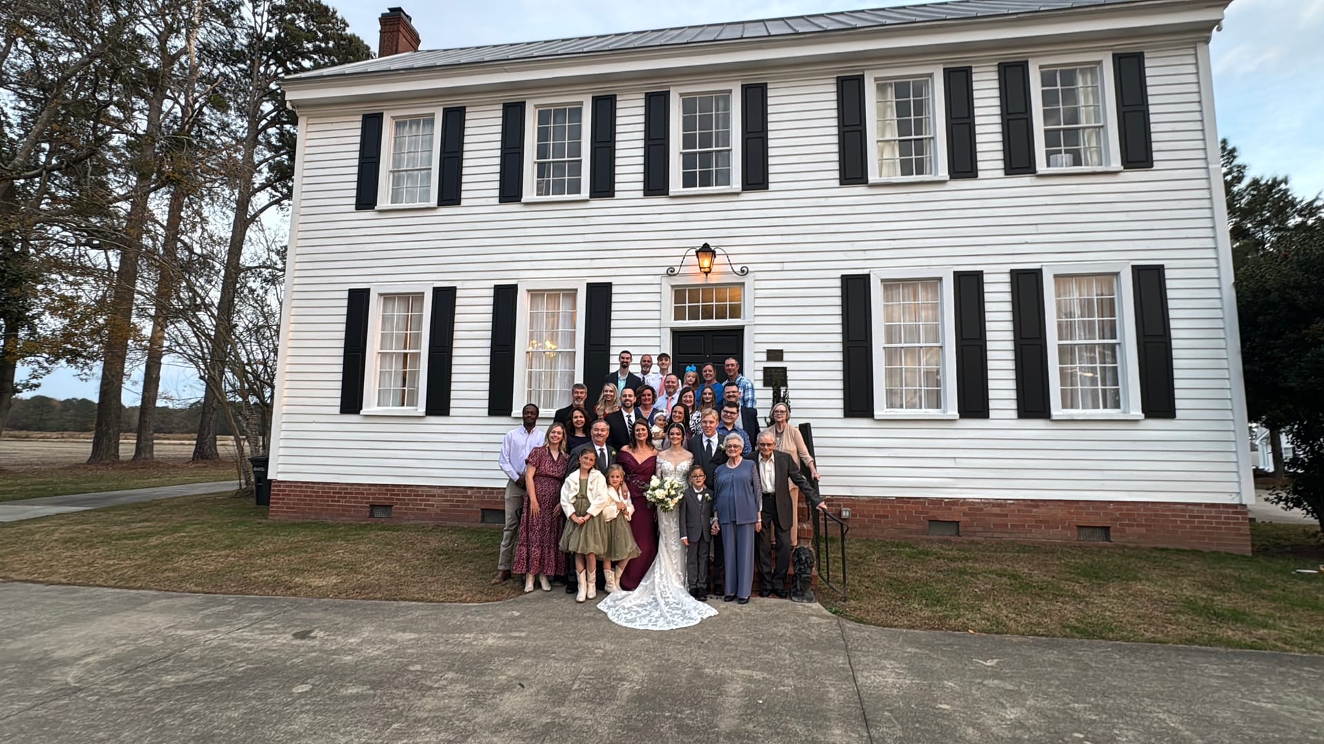 Wedding party and family gathered in front of the white 1821 Scarborough House manor with black shutters for a group portrait with the bride in a lace gown and groom in a dark suit at center.
