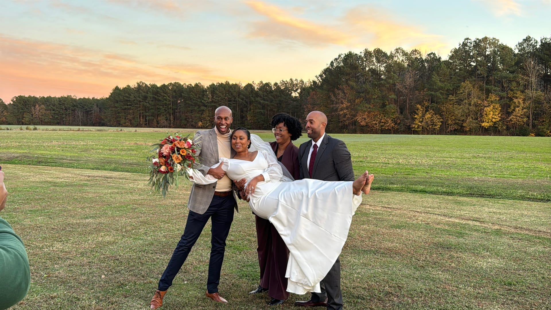 A joyful bride in a white gown holding an autumn bouquet is carried by two groomsmen while a bridesmaid in burgundy smiles beside them in an open farmland field at golden-hour sunset.