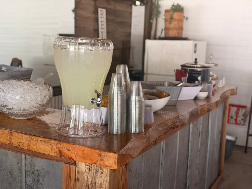 A large glass lemonade dispenser with plastic cups and a bowl of ice sits on a rustic live-edge wood bar at Scarborough House wedding venue.