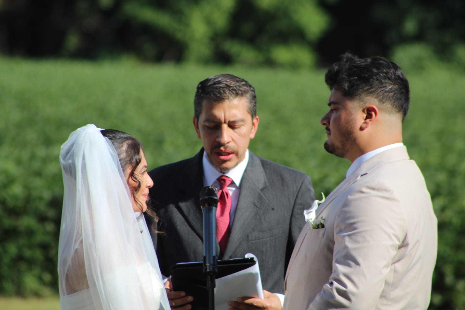A bride in a white veil and groom in a tan suit exchange vows before an officiant in a gray suit during an outdoor wedding ceremony surrounded by lush green fields and trees.