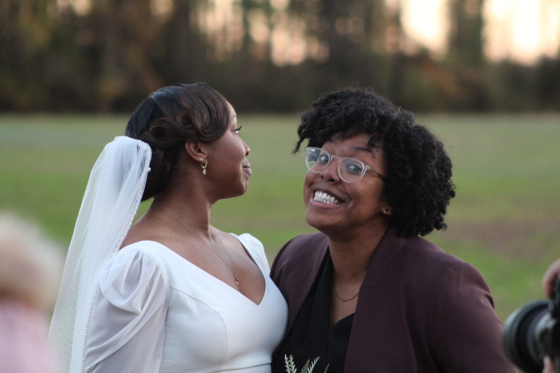 A smiling bride in a white gown and veil shares a joyful candid moment with a woman in a maroon blazer and glasses against a soft golden-hour field backdrop at Scarborough House.