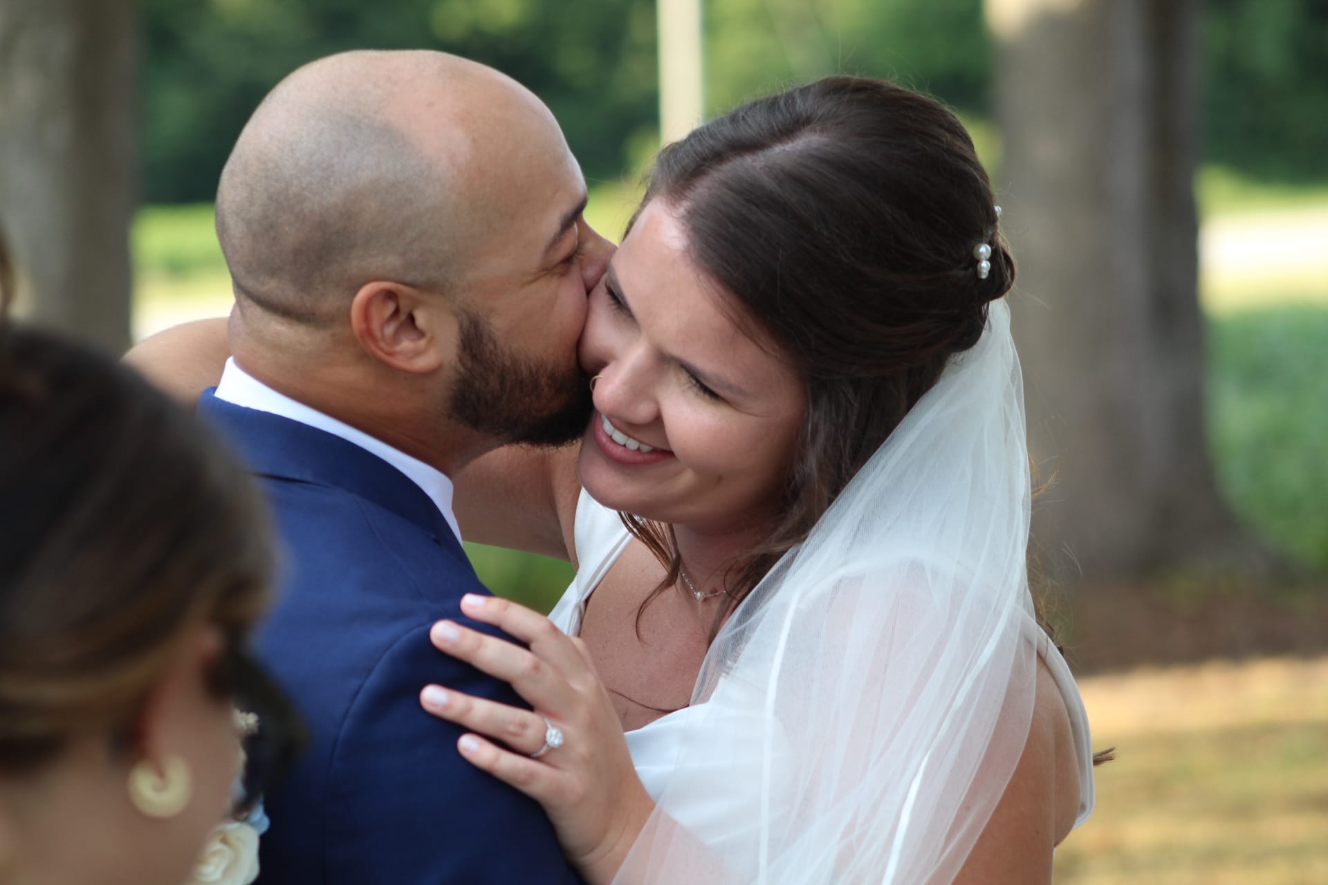 Groom in a navy suit kisses his smiling bride on the cheek beneath the oak trees at Scarborough House as she holds his arm, her white veil and diamond ring glowing in soft natural light.