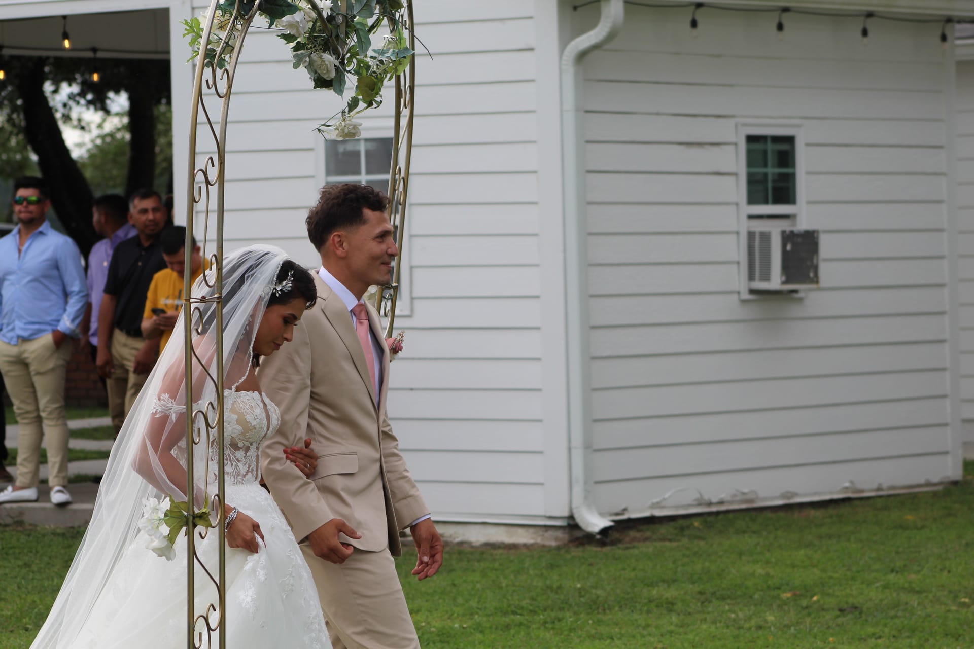 Bride in lace gown and veil walks arm in arm with groom in tan suit and pink tie through a gold floral arch on the ceremony lawn at Scarborough House.