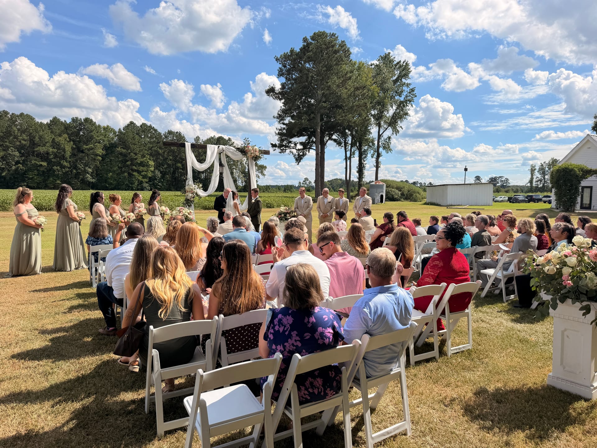 Guests seated in white folding chairs watch a sunny outdoor wedding ceremony on the ceremony lawn at Scarborough House, with a draped floral arch, bridesmaids in sage green, and open farmland fields in the background.