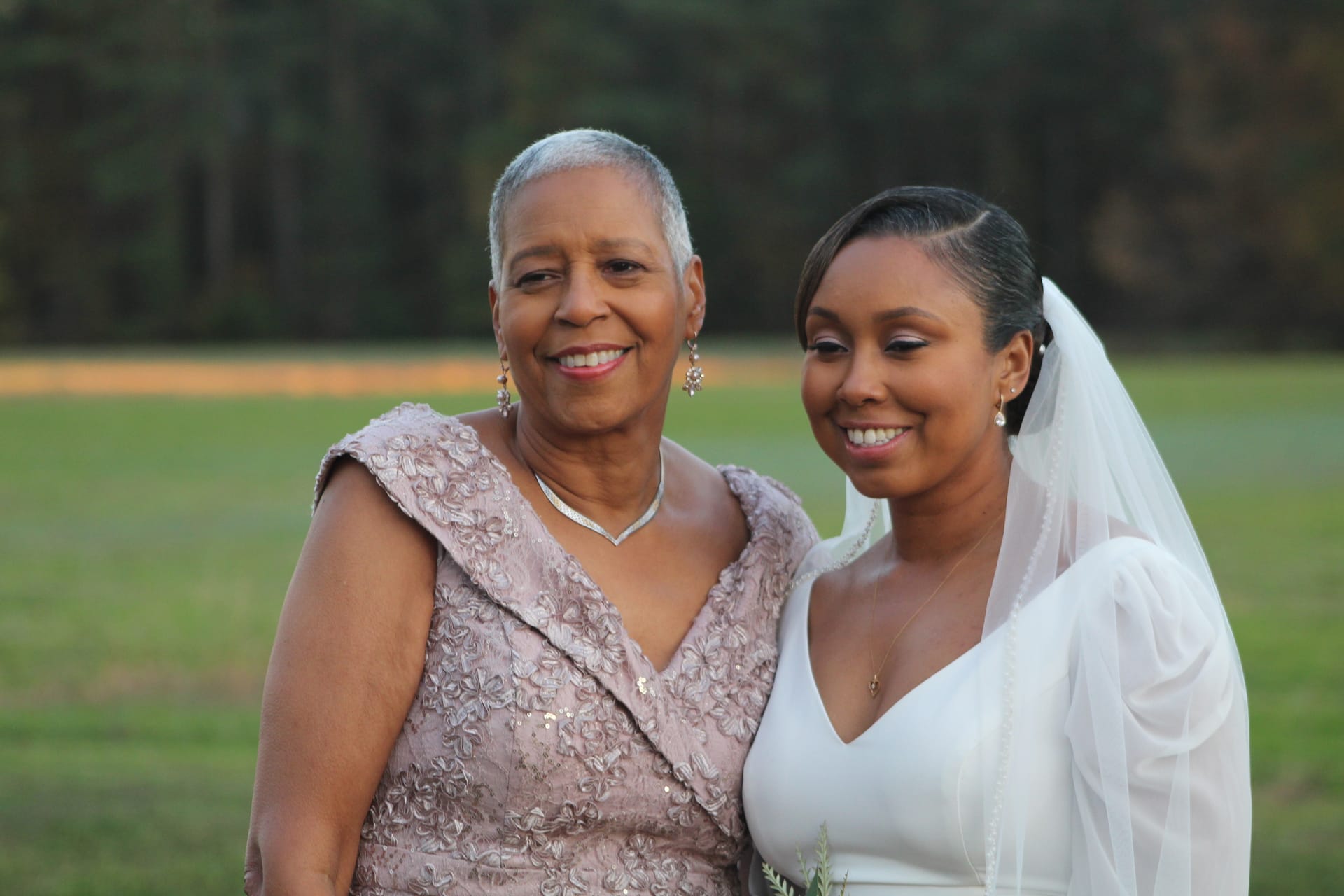 A smiling bride in a white gown and veil poses with an elegantly dressed older woman against a sunlit open field and tree line at Scarborough House.