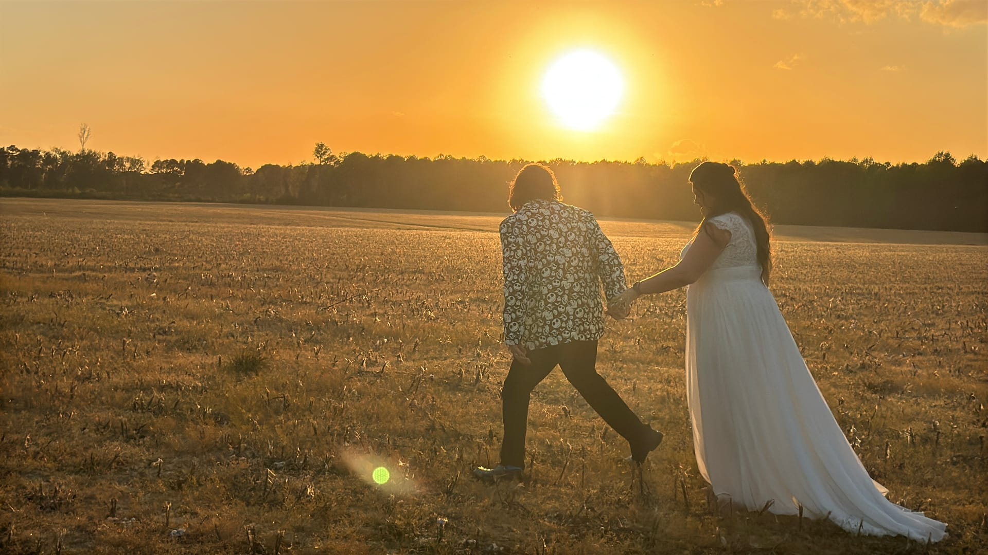 Bride in a flowing white gown and groom in a patterned shirt walk hand-in-hand across golden farmland fields at Scarborough House during a breathtaking sunset.