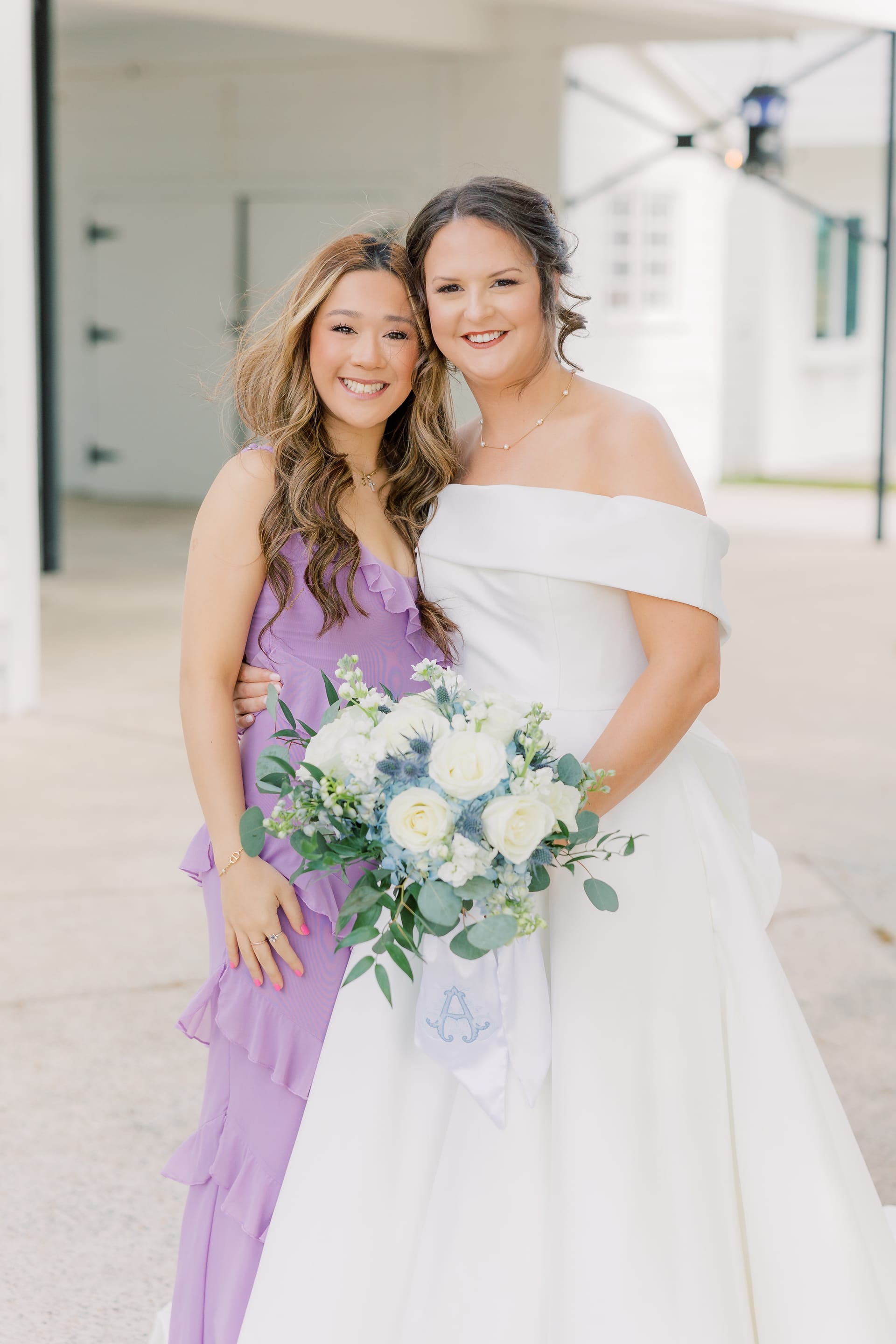 Bride in an off-the-shoulder white ball gown holding a white rose and eucalyptus bouquet poses with a smiling guest in a purple ruffle jumpsuit at Scarborough House.