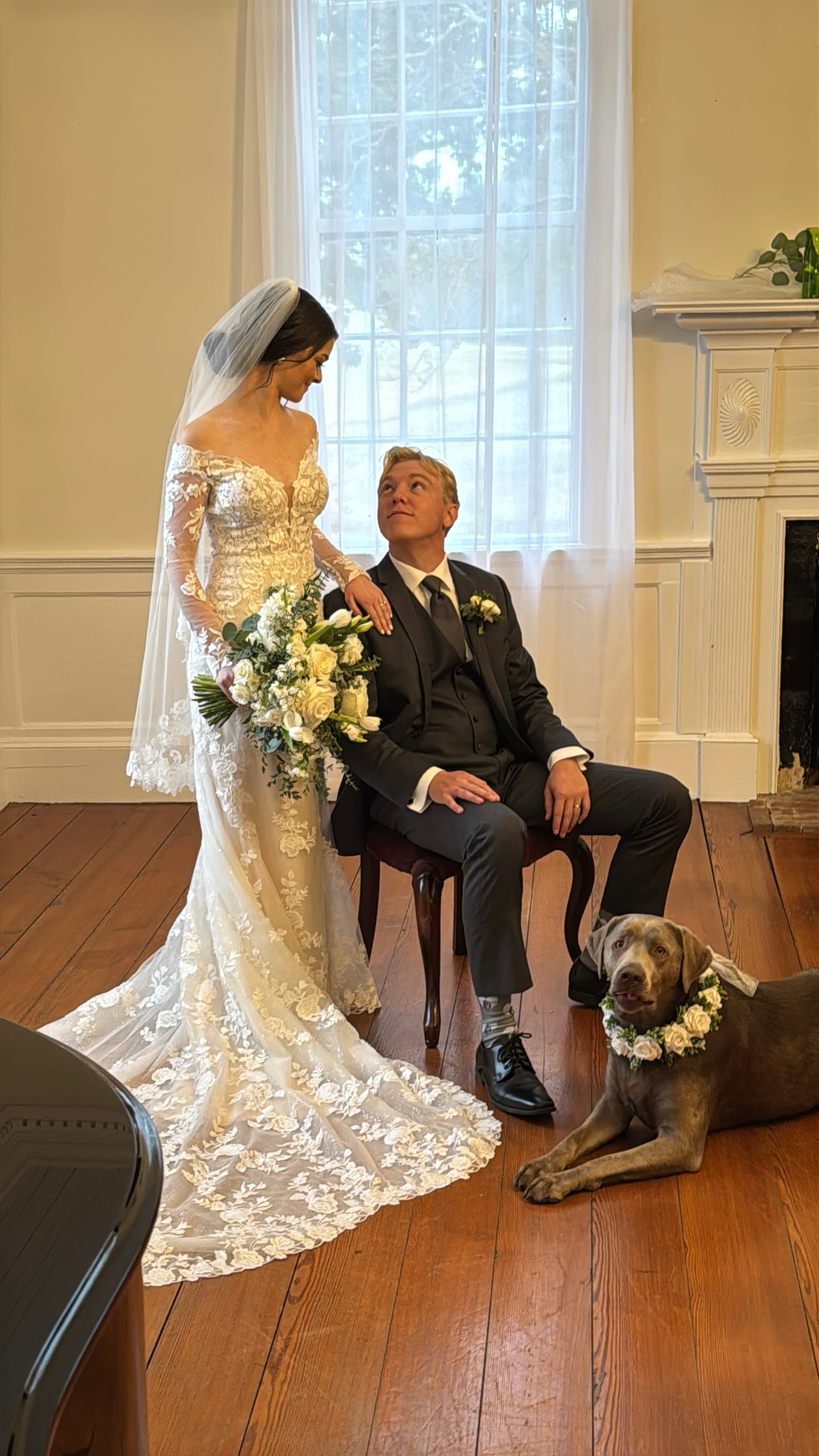 Bride in a lace off-shoulder gown holding a white floral bouquet stands beside her seated groom in a dark suit inside the historic 1821 manor grand hall, with their silver Labrador wearing a floral collar resting at their feet.