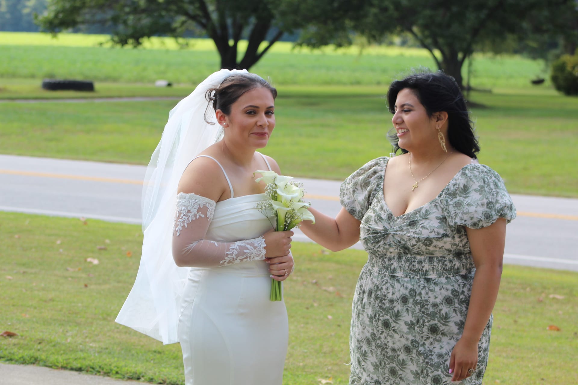 Bride in a fitted white gown with lace sleeves and veil holds a calla lily bouquet while a smiling companion in a floral dress adjusts her dress on the grounds of a historic estate wedding venue.