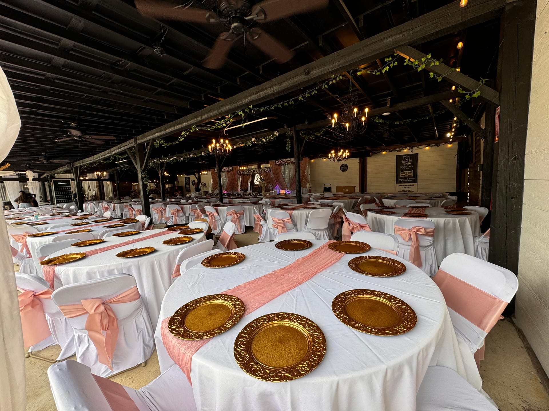 Elegantly decorated reception tables with white linens, blush pink sashes on chairs, gold ornate charger plates, and warm chandelier lighting inside the Magnolia Pavilion at Scarborough House.