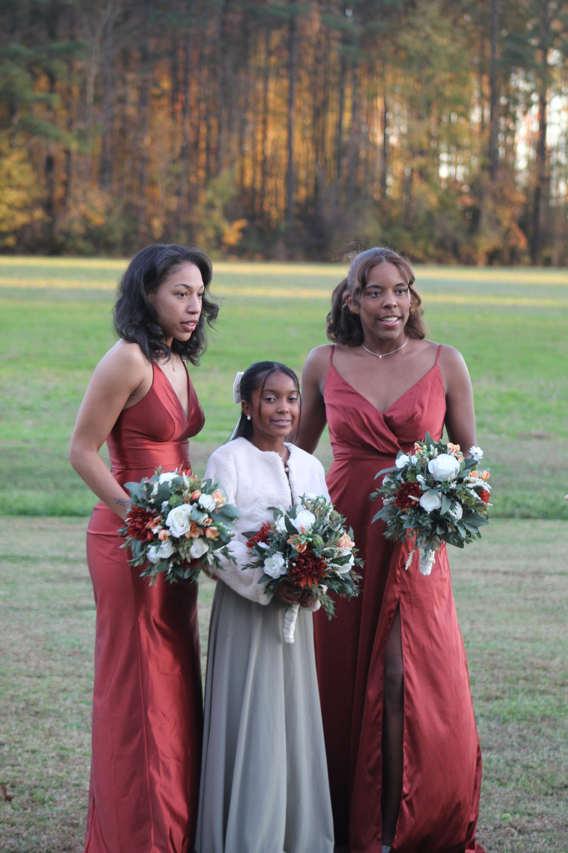 Two bridesmaids in burgundy satin gowns holding floral bouquets stand alongside a younger attendant in a white fur jacket on the open lawn at Scarborough House, surrounded by autumn trees.