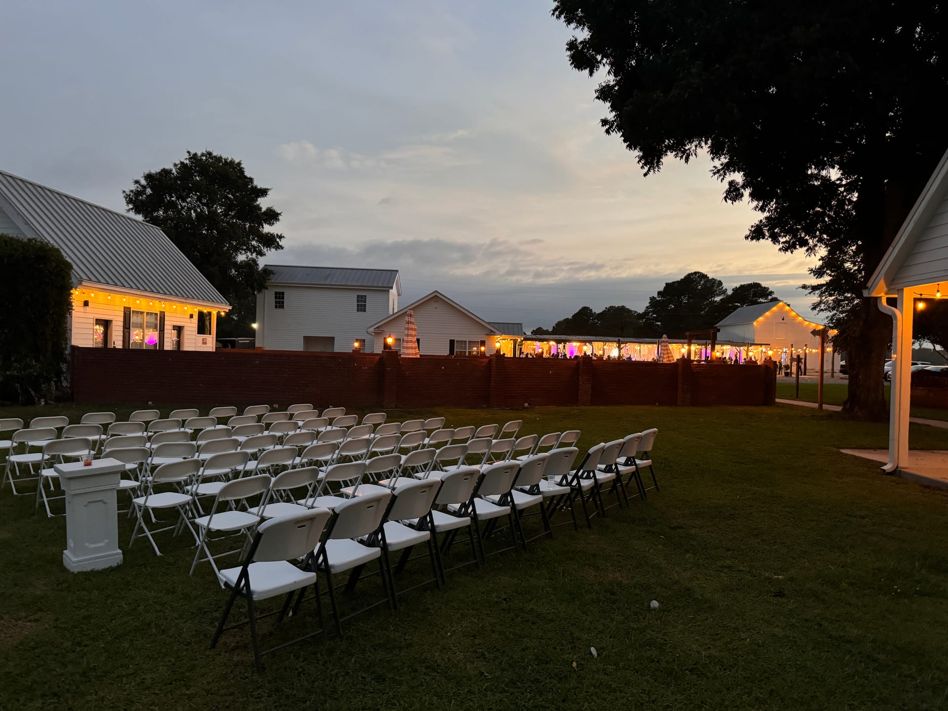 Rows of white folding chairs arranged on the ceremony lawn at dusk with string lights and illuminated venue buildings glowing warmly in the background at Scarborough House.