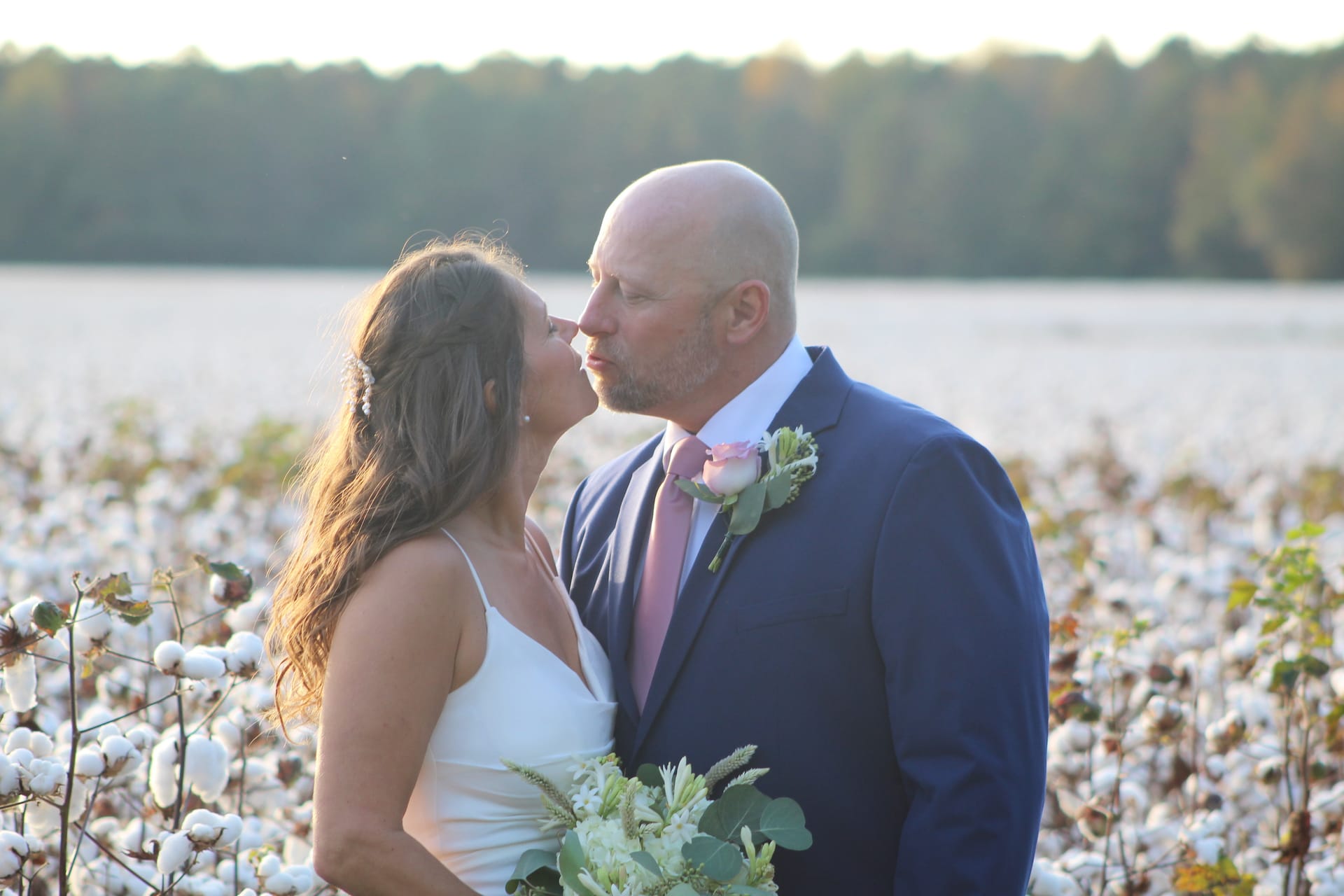 Bride and groom sharing a tender almost-kiss in a sunlit cotton field at golden hour, surrounded by blooming cotton bolls with a treeline glowing in the background.