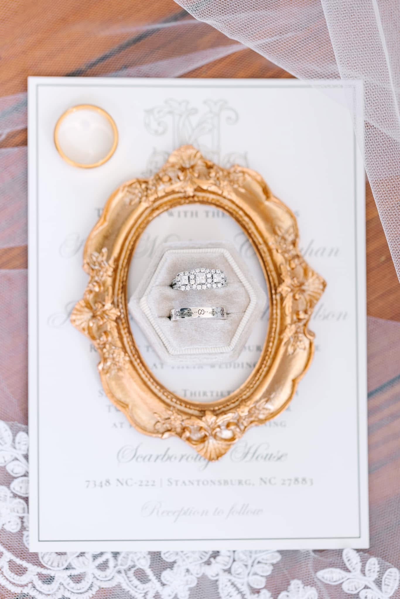 Wedding rings displayed in a cream velvet hexagon ring box atop an ornate gold oval frame and printed Scarborough House wedding invitation, accented by a gold band and bridal veil.