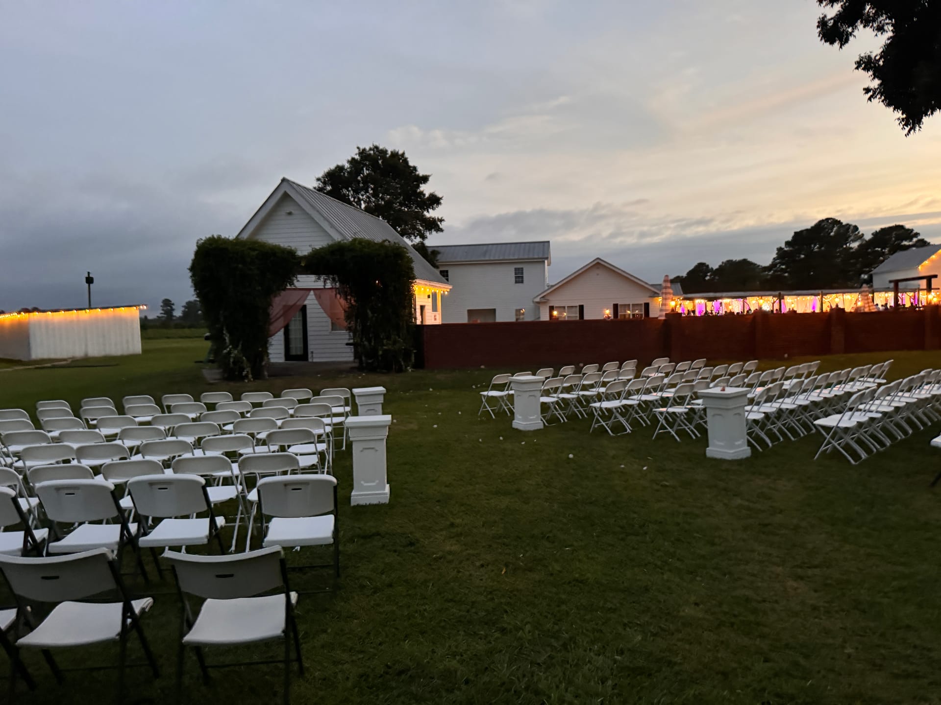 White folding chairs arranged in rows on the ceremony lawn at Scarborough House at dusk with warm string lights and the historic manor glowing softly in the background.