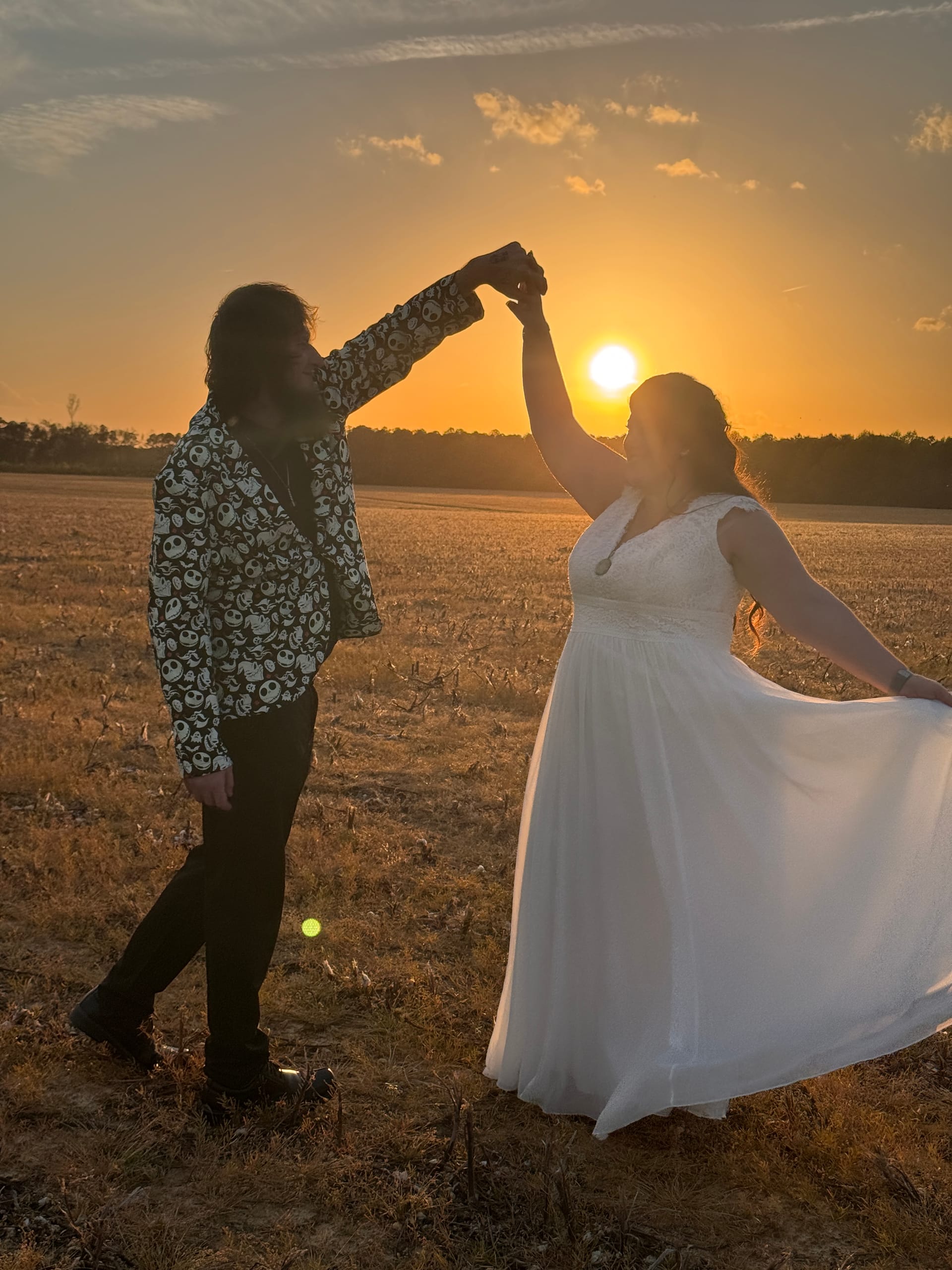 Bride in flowing white gown being twirled by groom in a patterned jacket as they dance in an open farmland field at sunset with a vivid orange sky behind them.