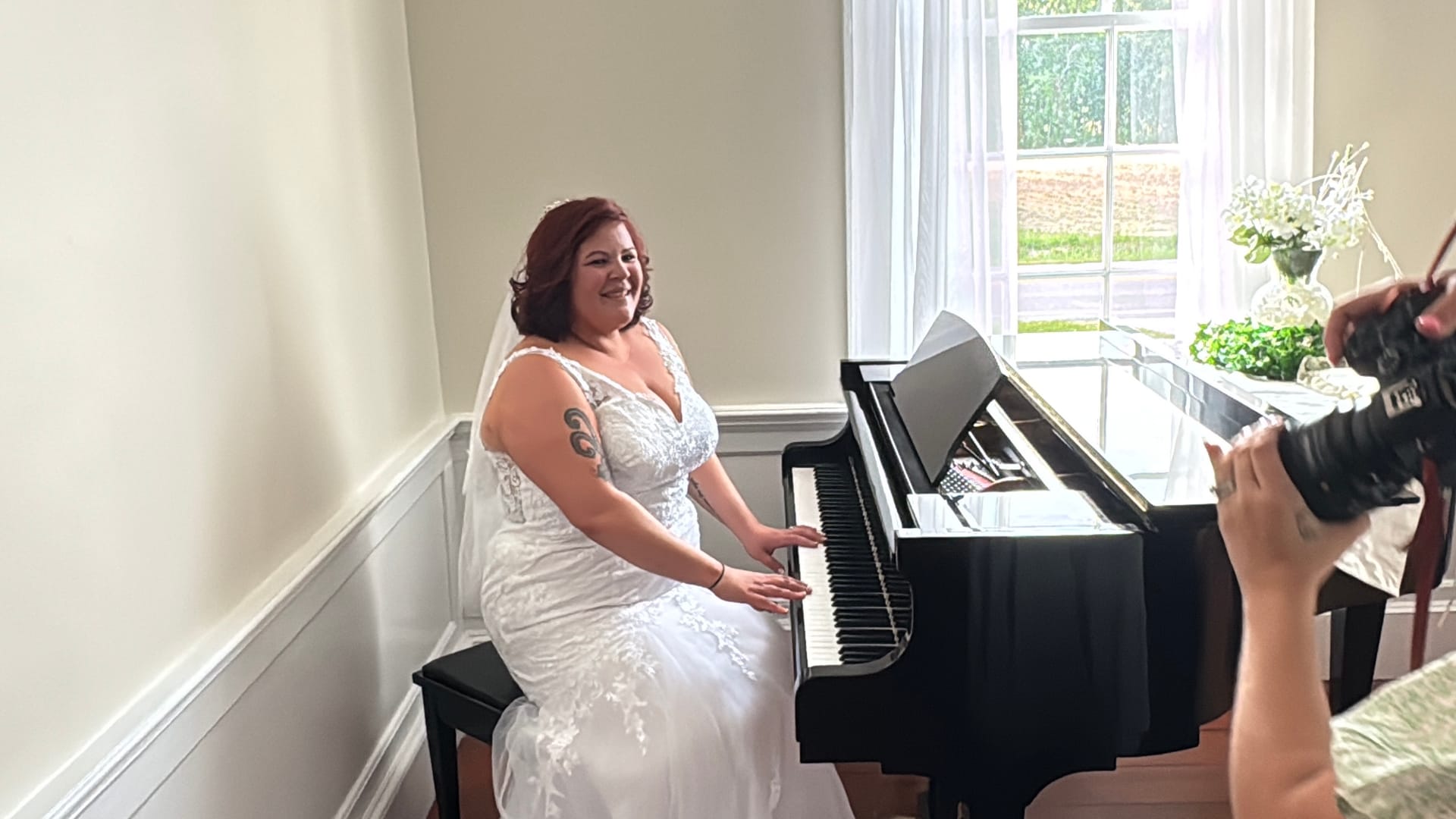 A bride in a white lace gown and veil smiles warmly while seated at a black baby grand piano near a bright window inside the historic Scarborough House manor.