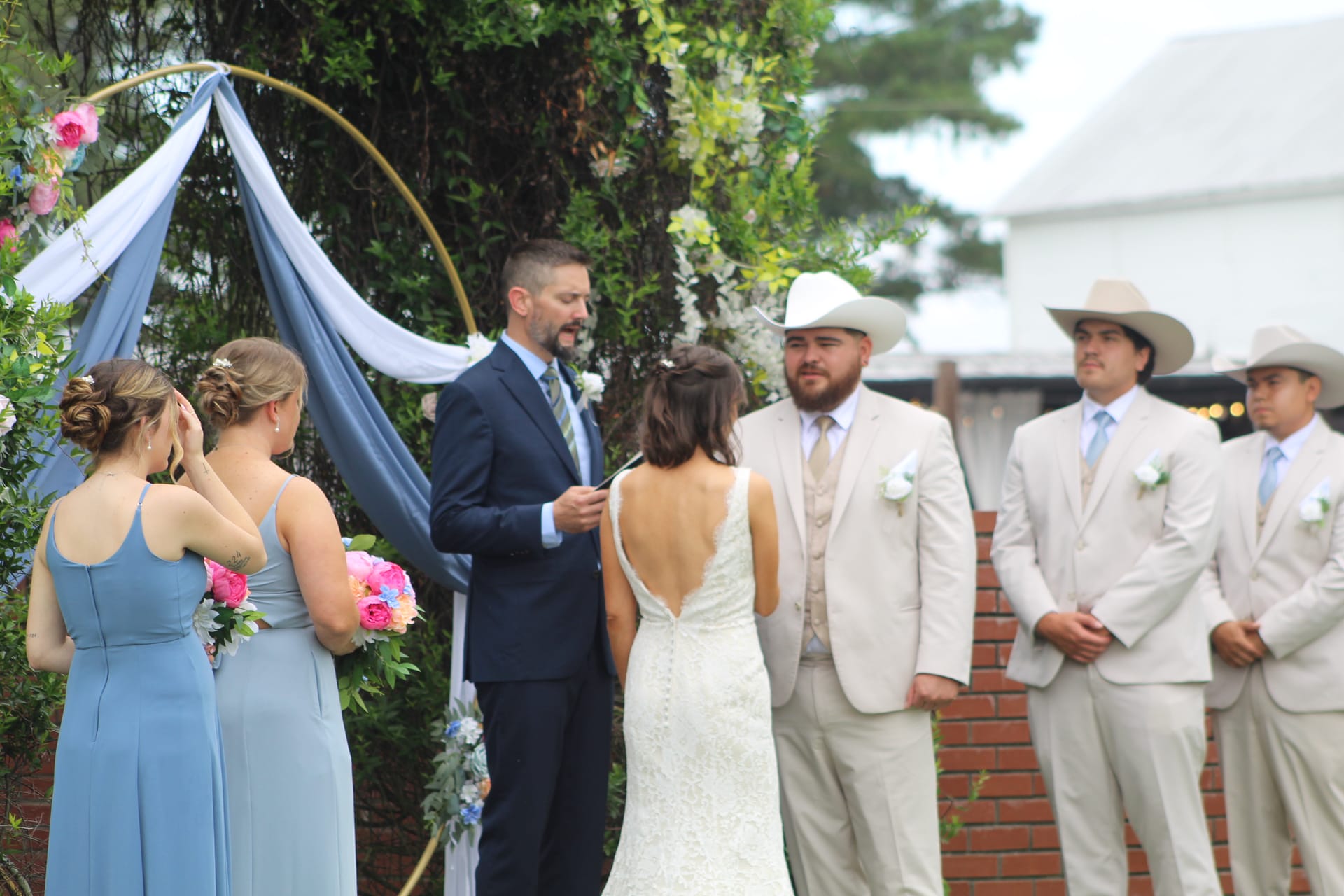 Bride in a backless lace gown stands before an officiant under a floral gold hoop arch with blue draping as emotional bridesmaids and cowboy-hat-wearing groomsmen look on during an outdoor wedding ceremony at Scarborough House.