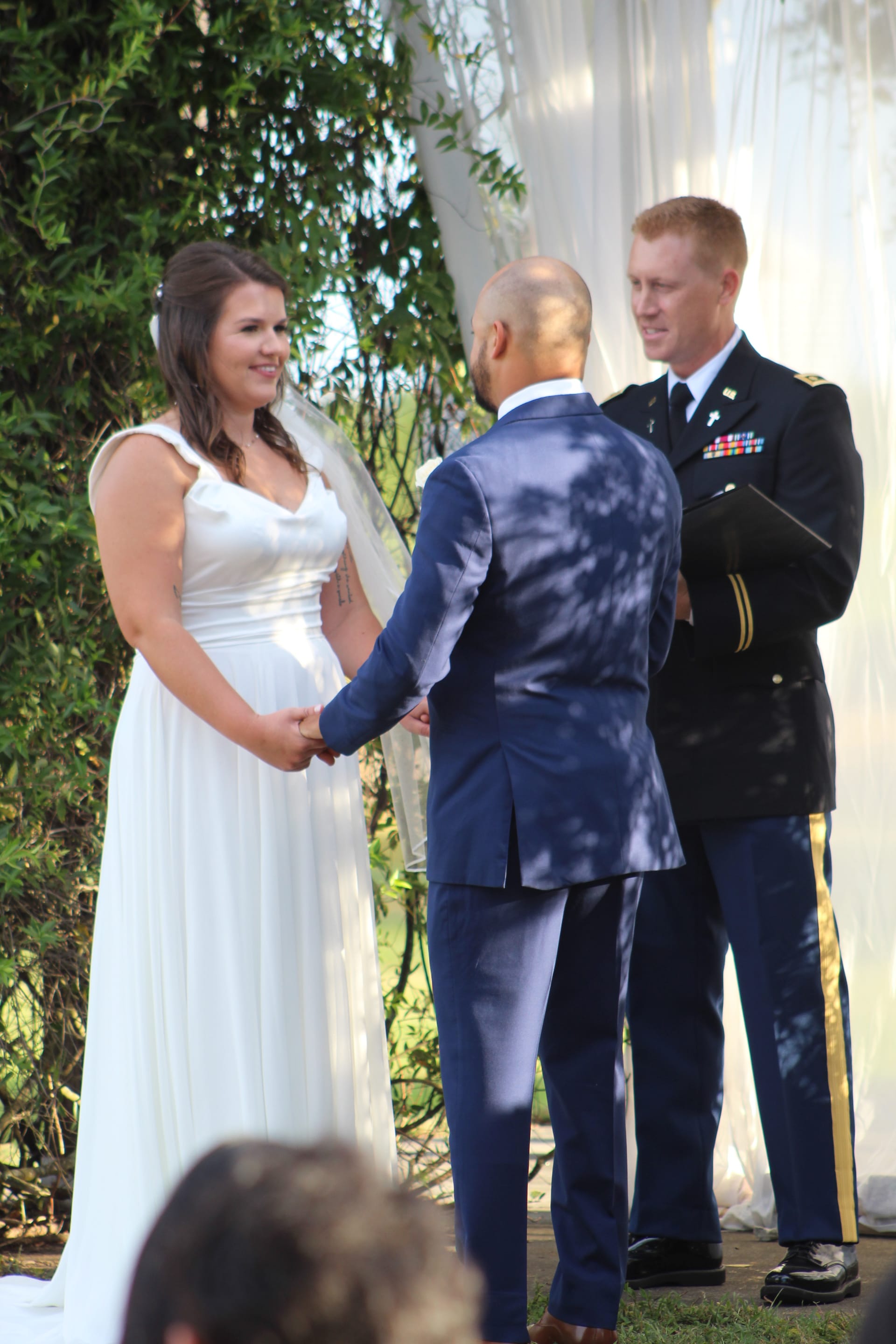 Bride in a flowing white gown smiles warmly at her groom as they hold hands during an outdoor wedding ceremony officiated by a military chaplain at Scarborough House.