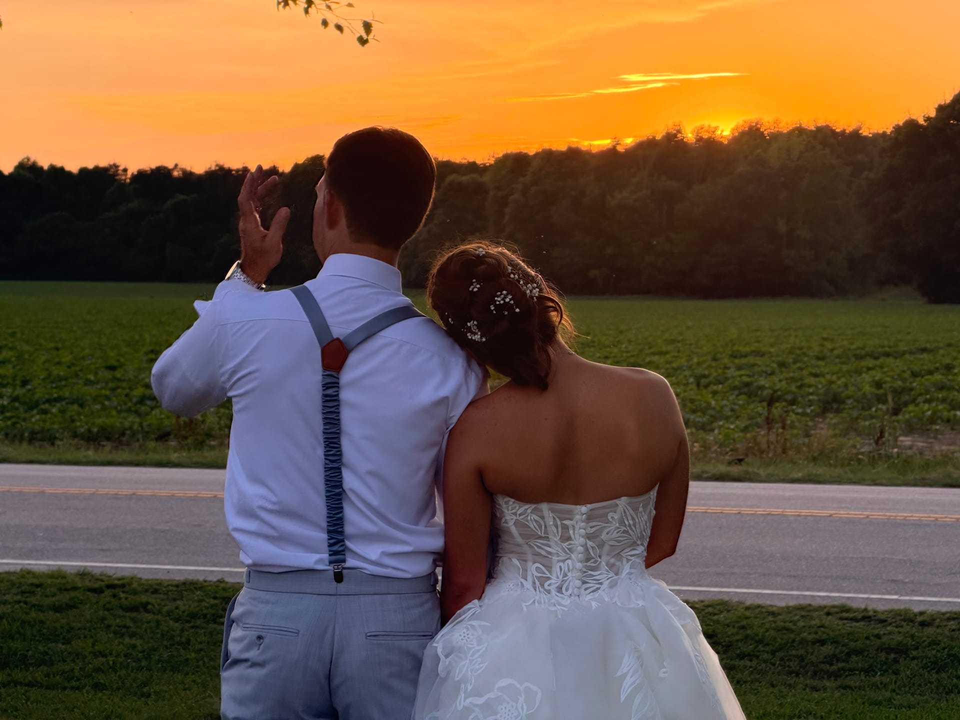 Bride in a strapless lace gown and groom in light grey suit with blue suspenders stand together facing a brilliant orange sunset over open farmland fields at Scarborough House.