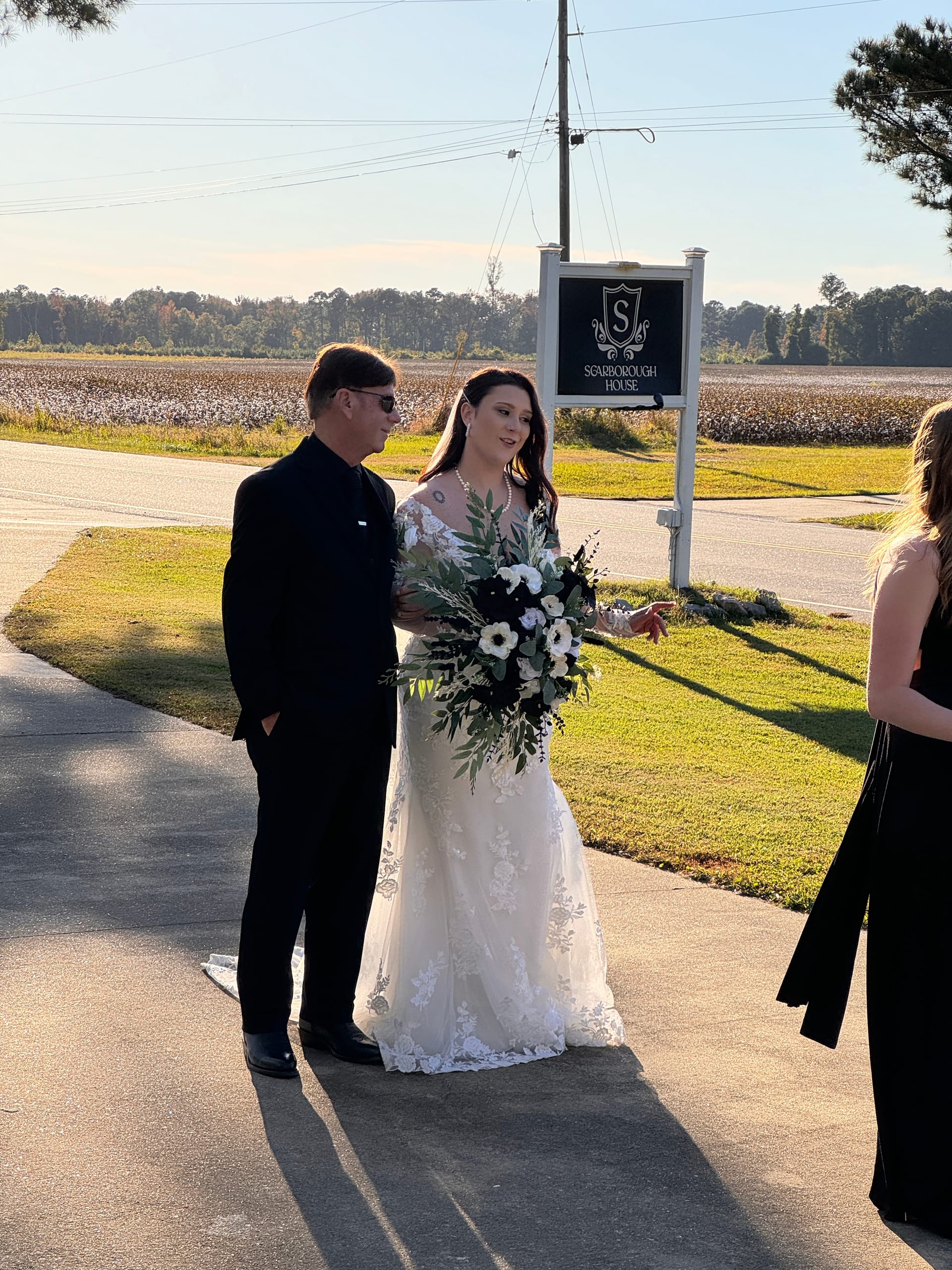 Bride in a floral lace wedding gown holding a cascading black and white bouquet stands with a gentleman in black beside the Scarborough House marquee sign, with cotton fields stretching out behind them on a sunny autumn day.