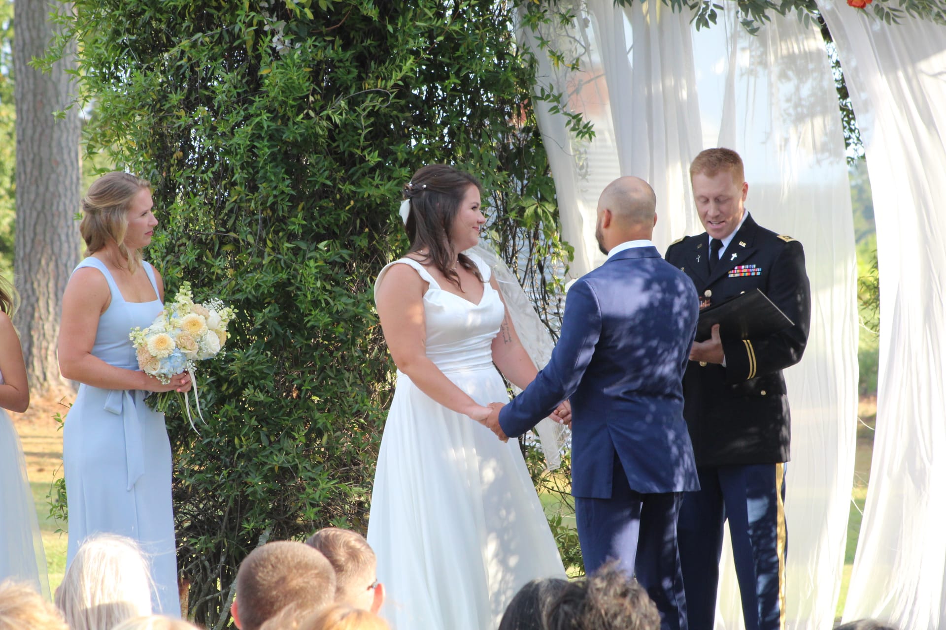 Bride in a white gown smiles while exchanging vows with her groom before a military officiant beneath a draped floral arch on the sunlit ceremony lawn at Scarborough House.